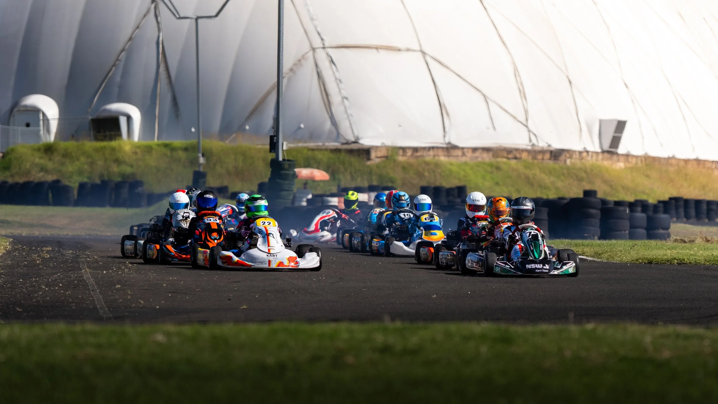 Go-kart race with multiple racers on a track, all wearing helmets, with a large white aerodynamic structure in the background.
