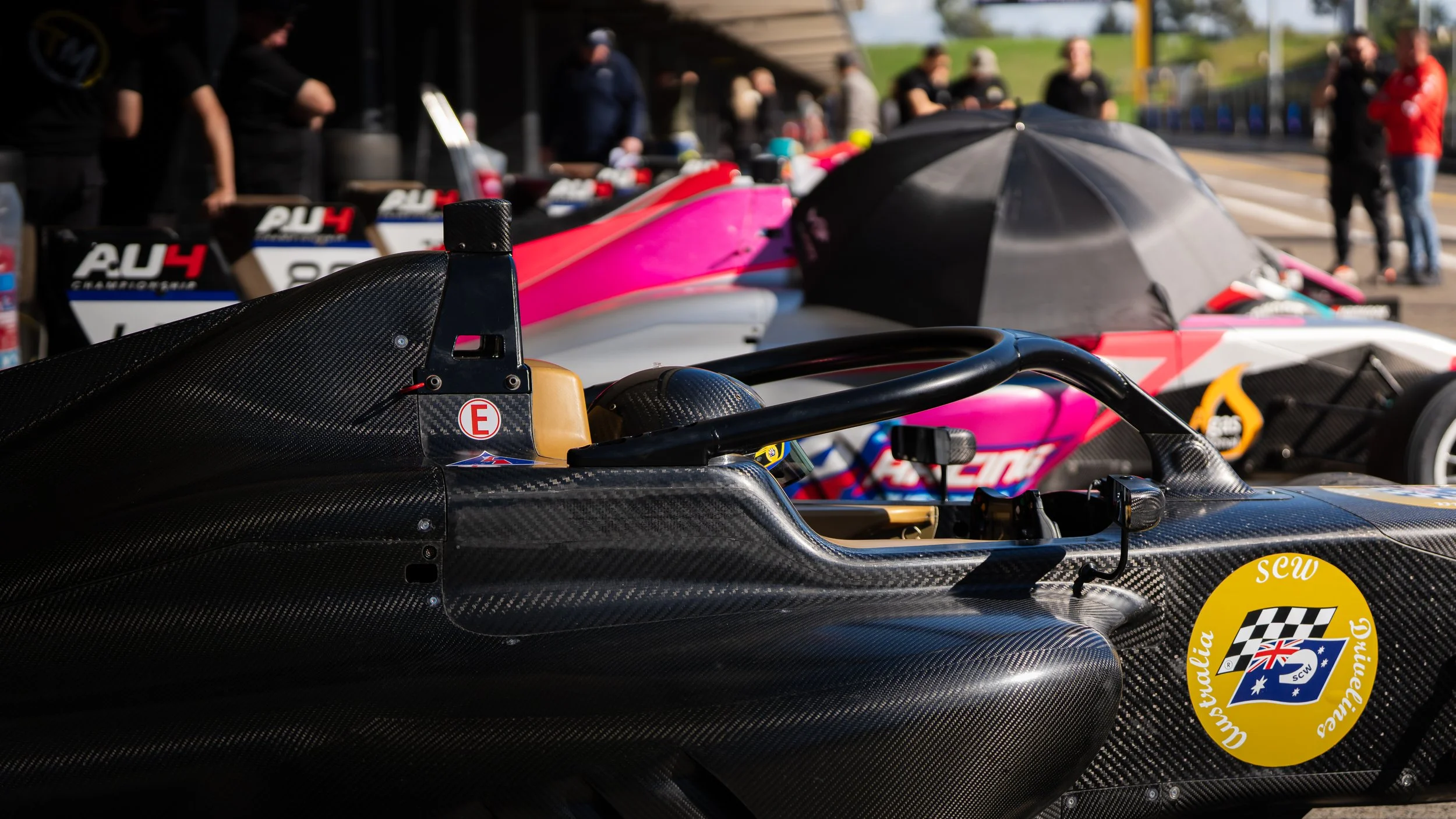 Close-up of a black racing car with a yellow sticker, showing a cockpit with a steering wheel and a carbon fiber body, in a racing pit area with pink and white cars and people in the background