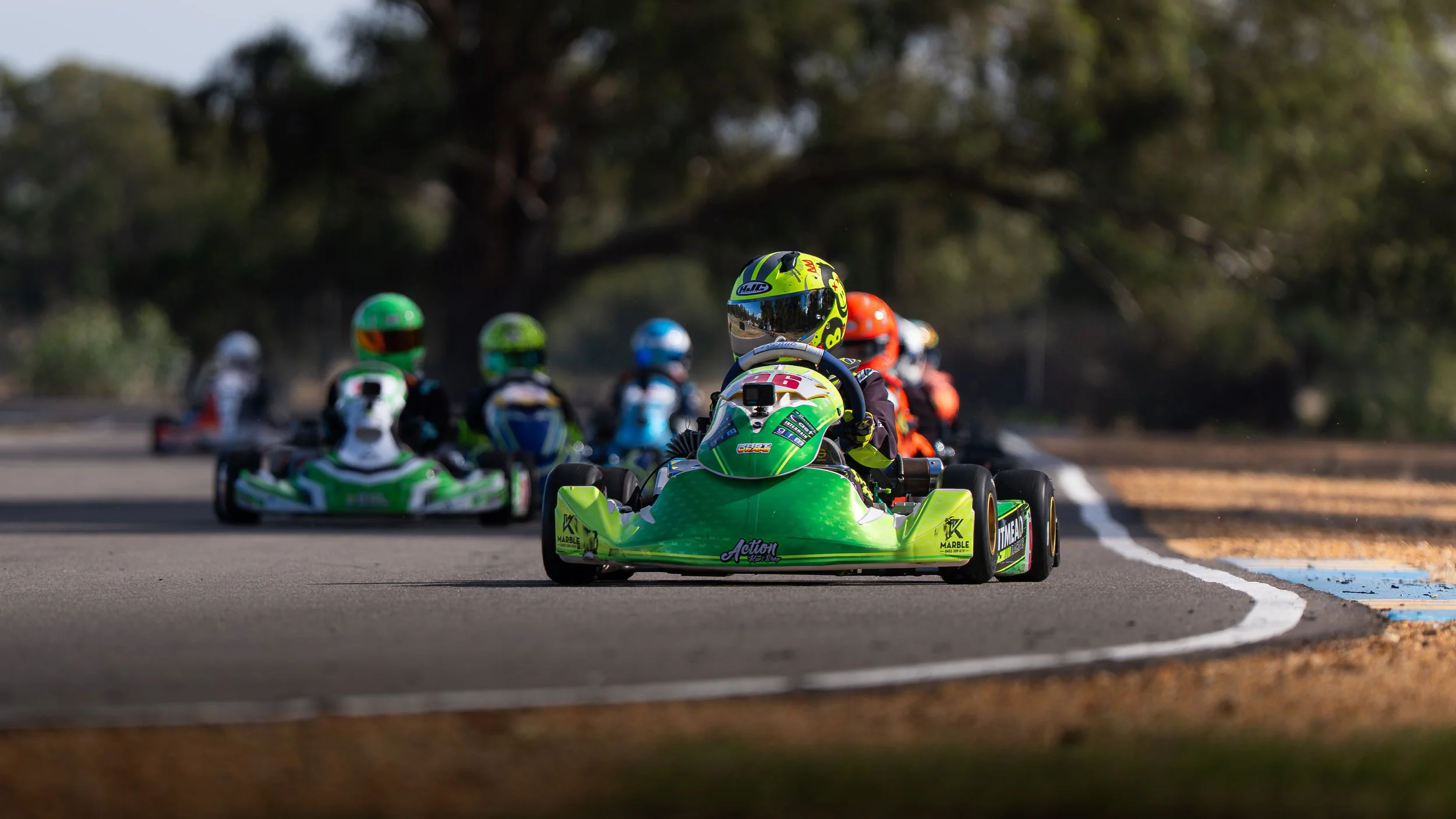Go-kart race with multiple racers wearing colorful helmets on a track, with trees in the background.