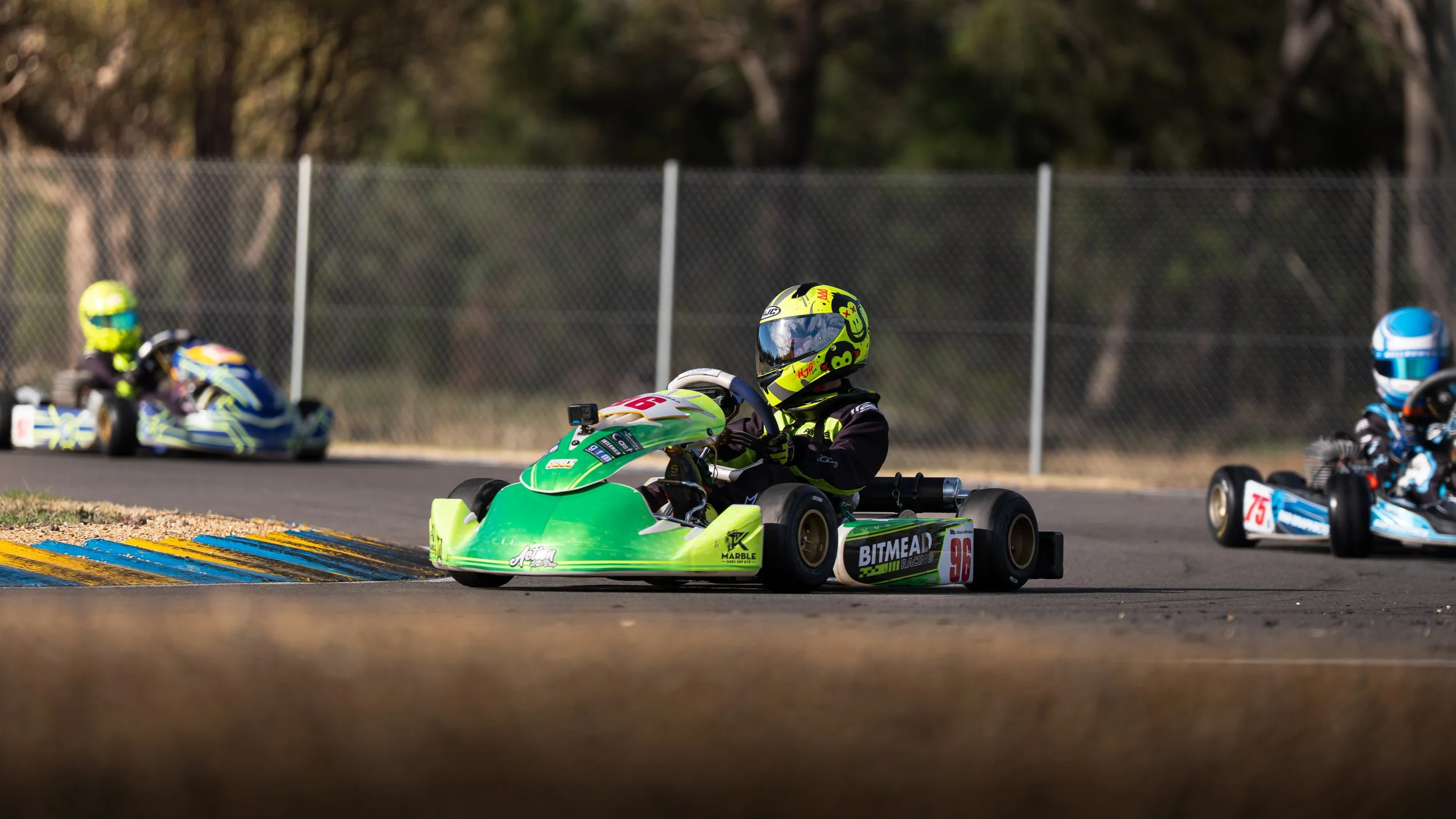 Three go-kart racers on a track, with one green go-kart in the foreground and two other racers in the background, all wearing helmets and racing suits.