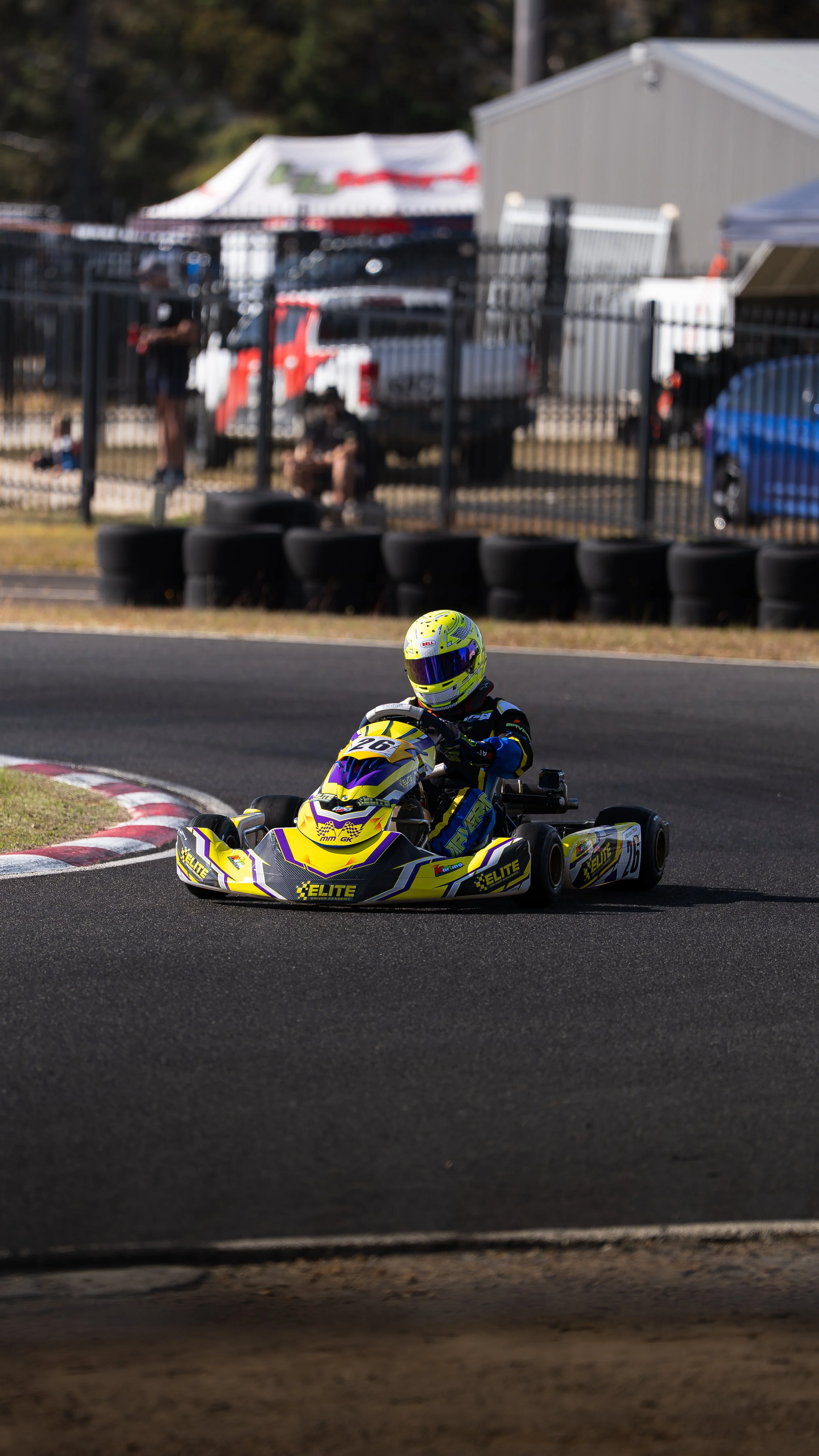 A go-kart racer in a yellow and purple suit and helmet driving on a race track with black tires and a fence in the background.
