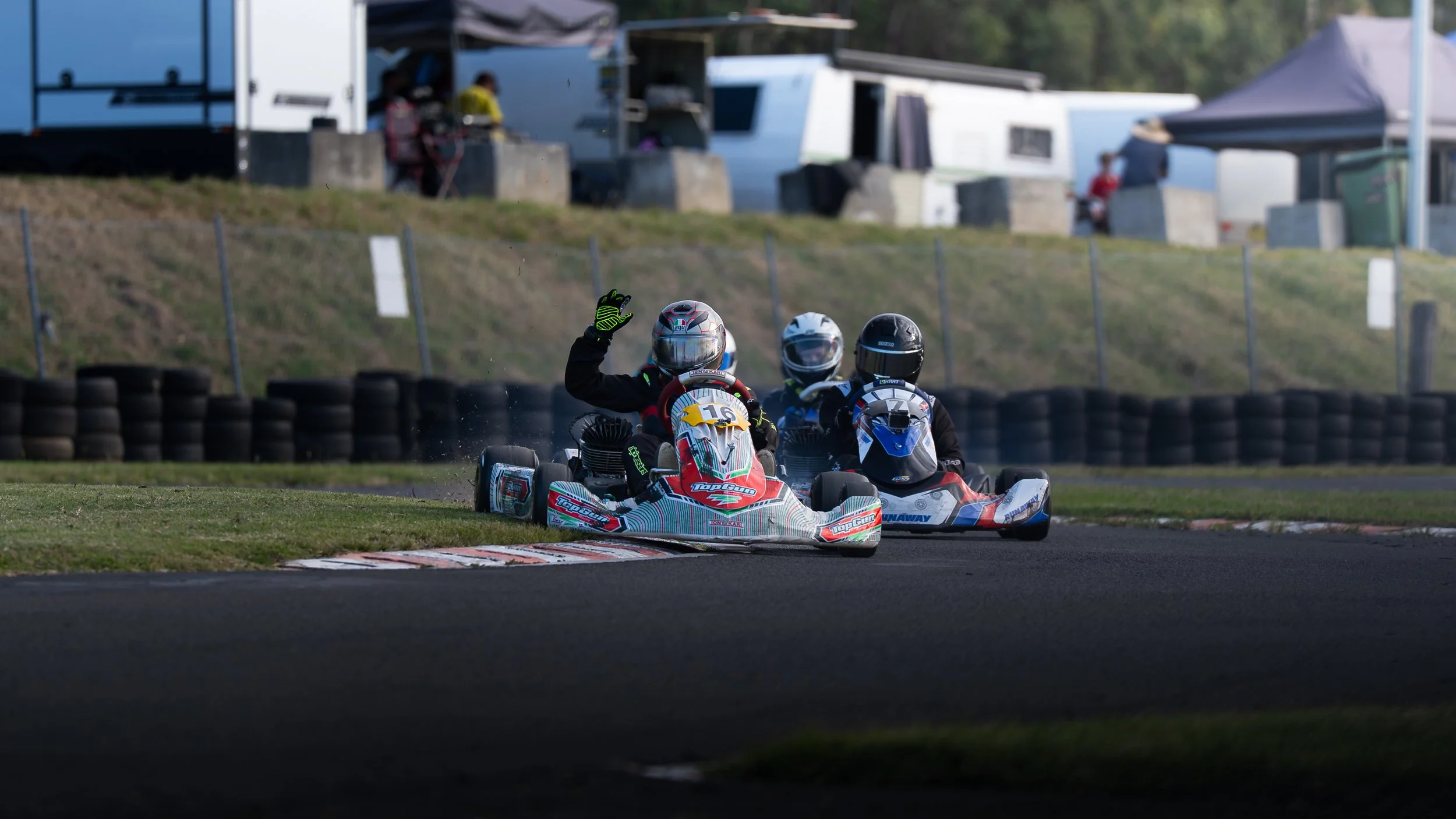 Two go-karts racing on a track with three racers wearing helmets, one waving, in an outdoor setting with tents and trailers in the background.