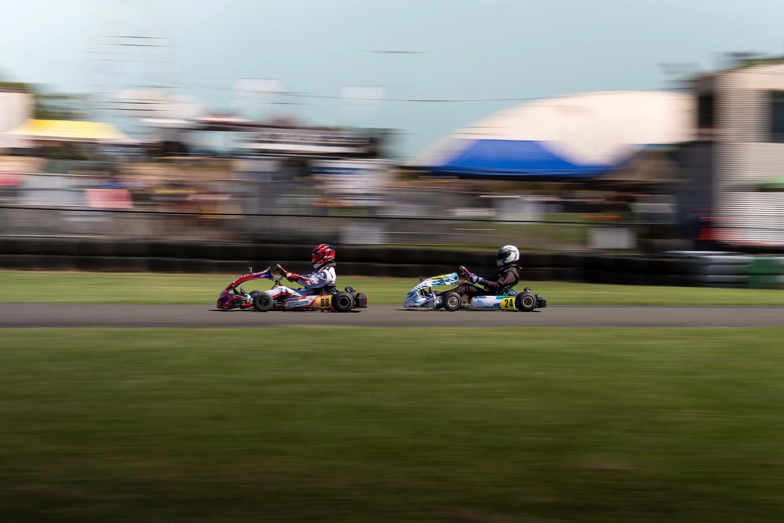 Two go-kart racers wearing helmets and racing suits on a track, with blurred background indicating motion.