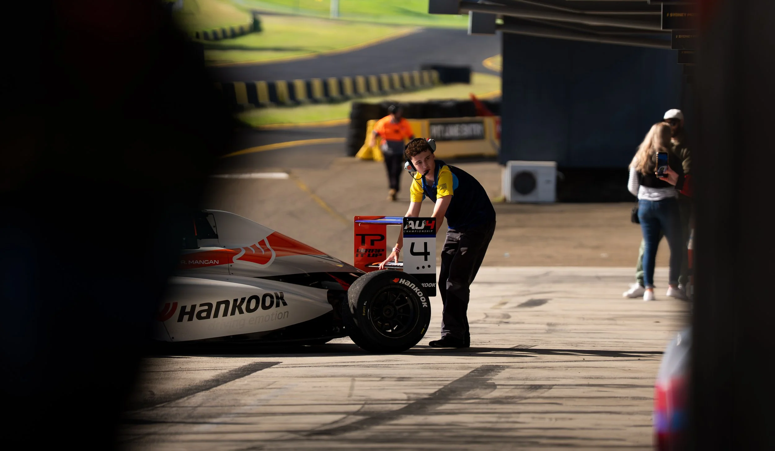A young race car driver in a racing suit and headphones standing beside a racing car in a pit area, with a woman taking a photo nearby and another person in the background.