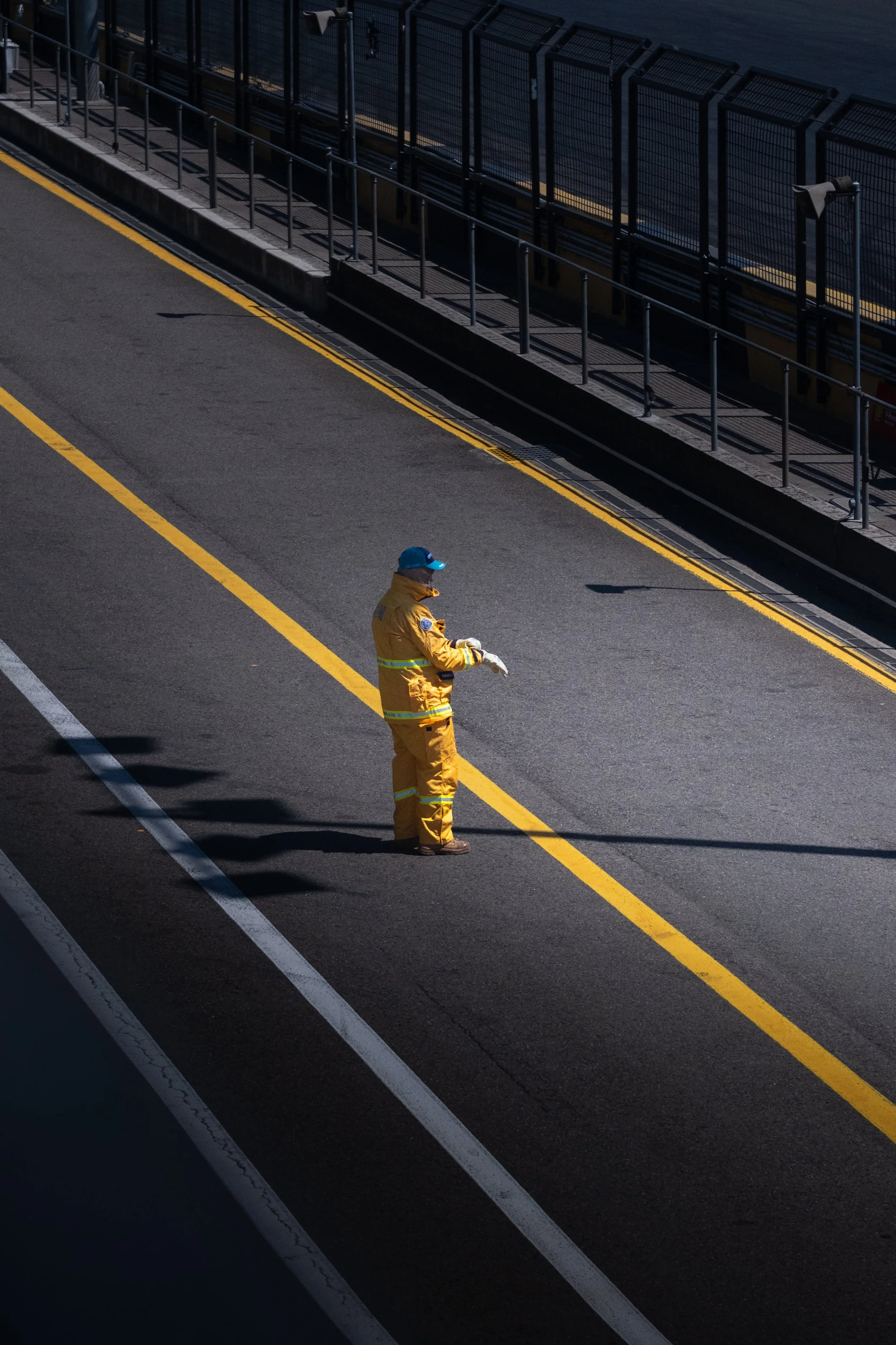 A person in yellow safety gear and a blue cap standing on a dark racetrack with yellow lines. There is a fence along the edge of the track with lighting fixtures.