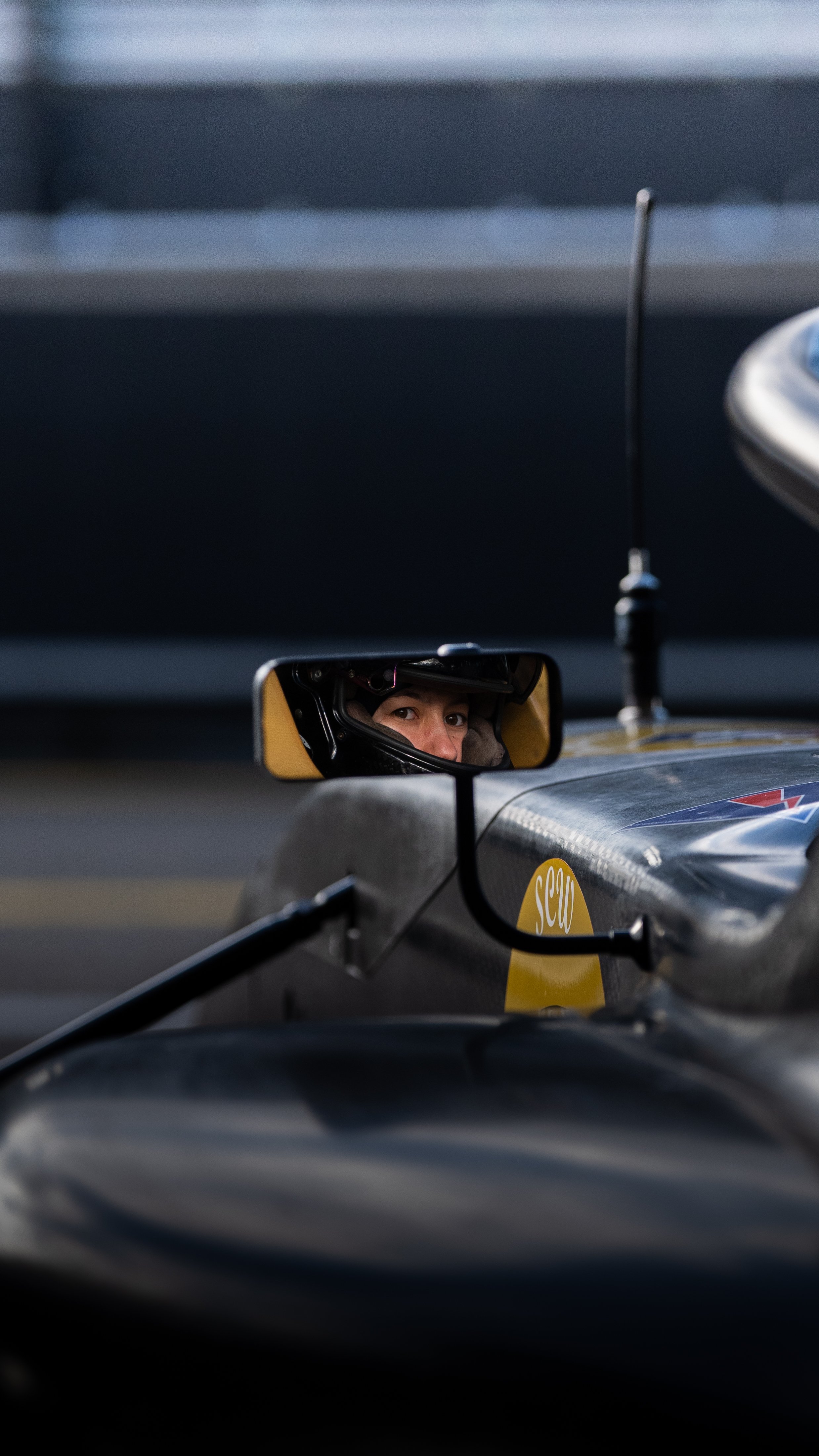 A race car driver is seen through the side mirror, wearing a helmet and looking at the camera inside a racing car.
