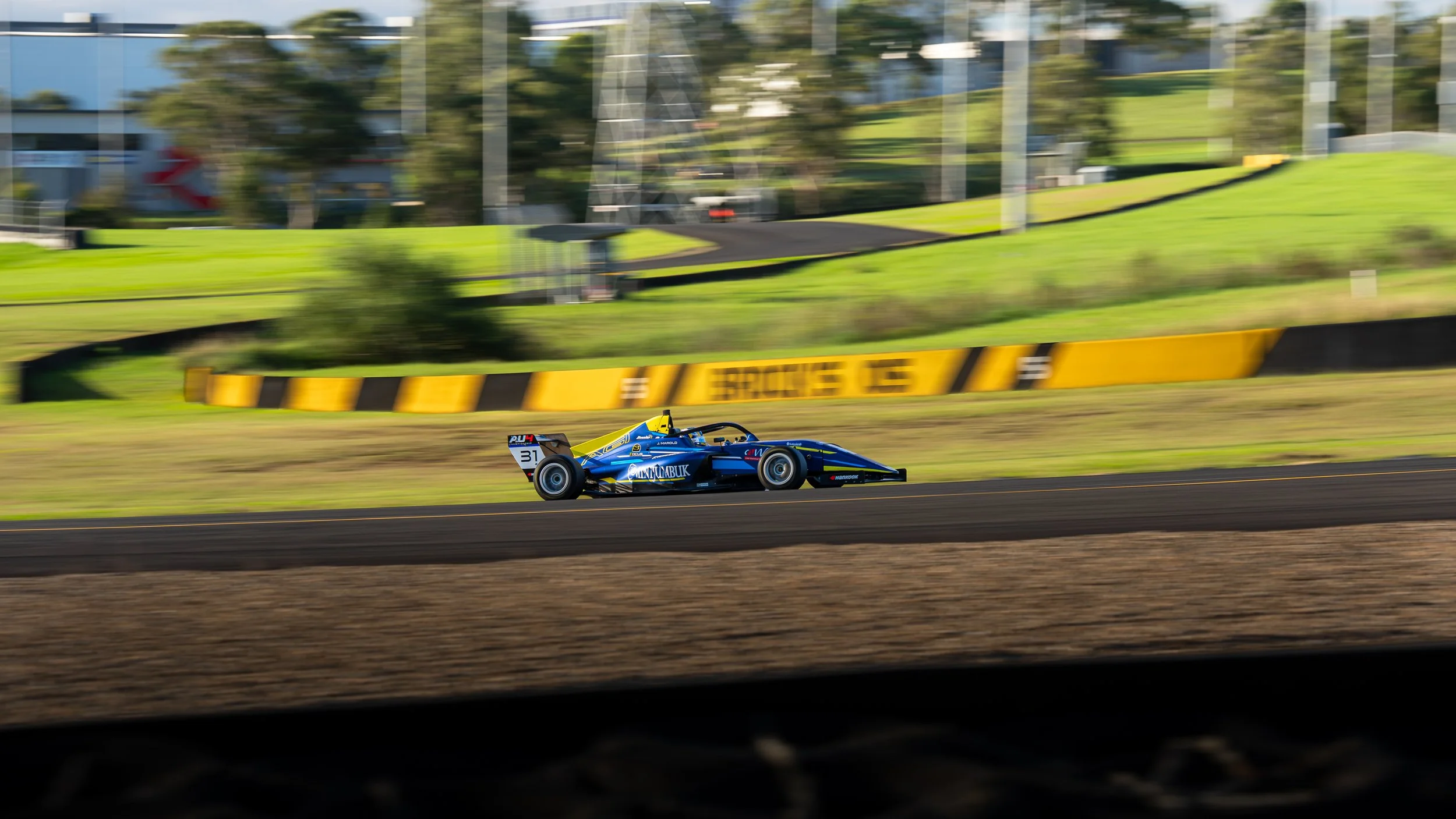 A race car traveling on a racetrack, with a motion blur effect showing speed, surrounded by green grass and some structures in the background.