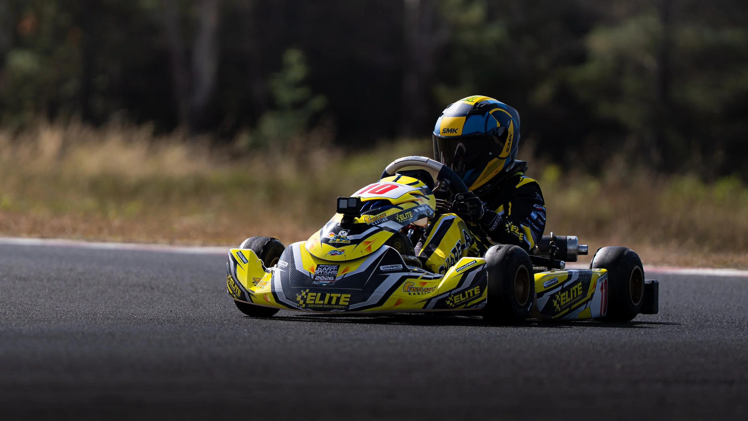 A person racing in a yellow go-kart on a track, wearing a helmet, surrounded by a blurred background of trees.