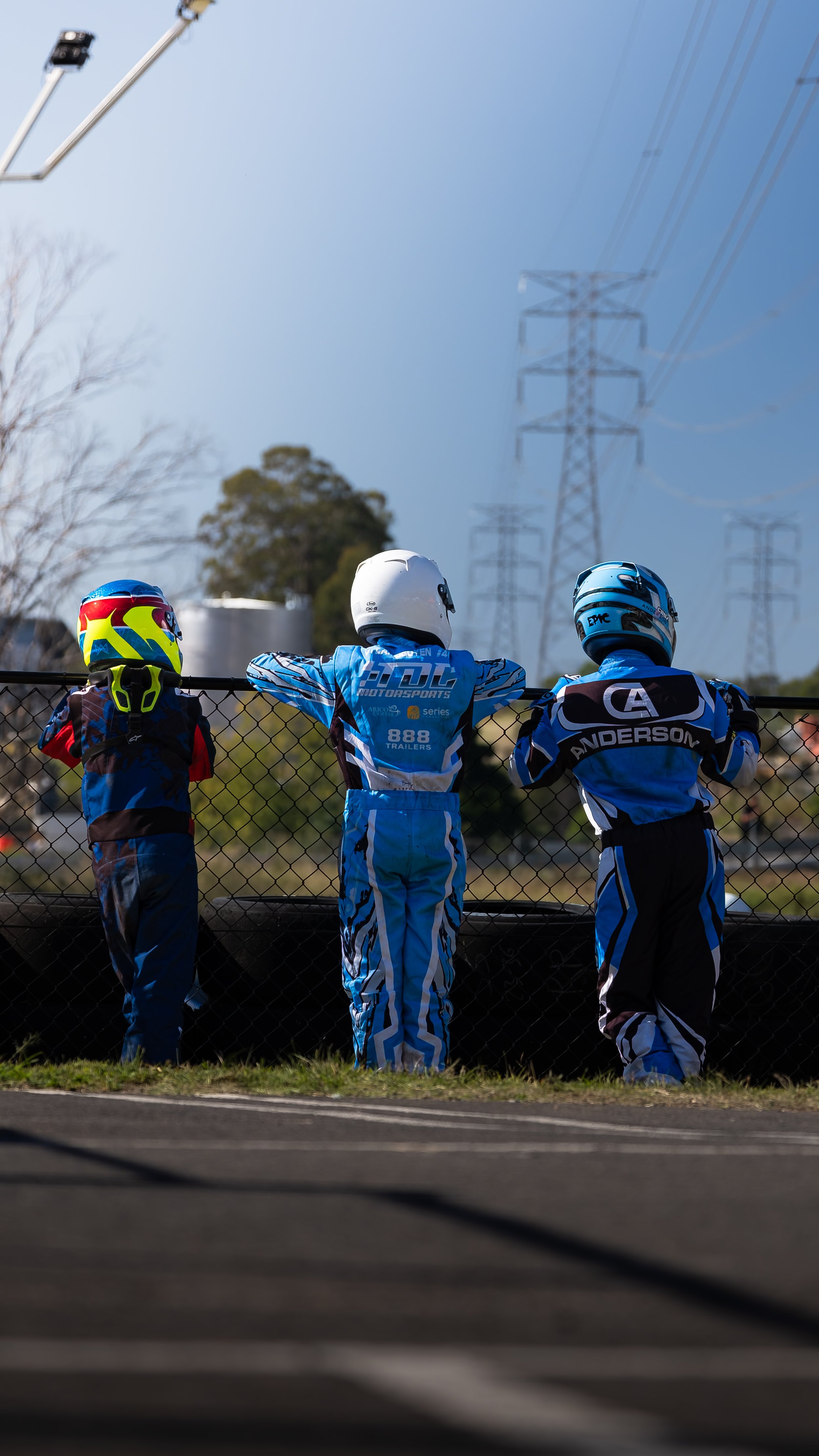 Three young race car drivers in racing suits and helmets standing behind a racetrack fence, watching the race.