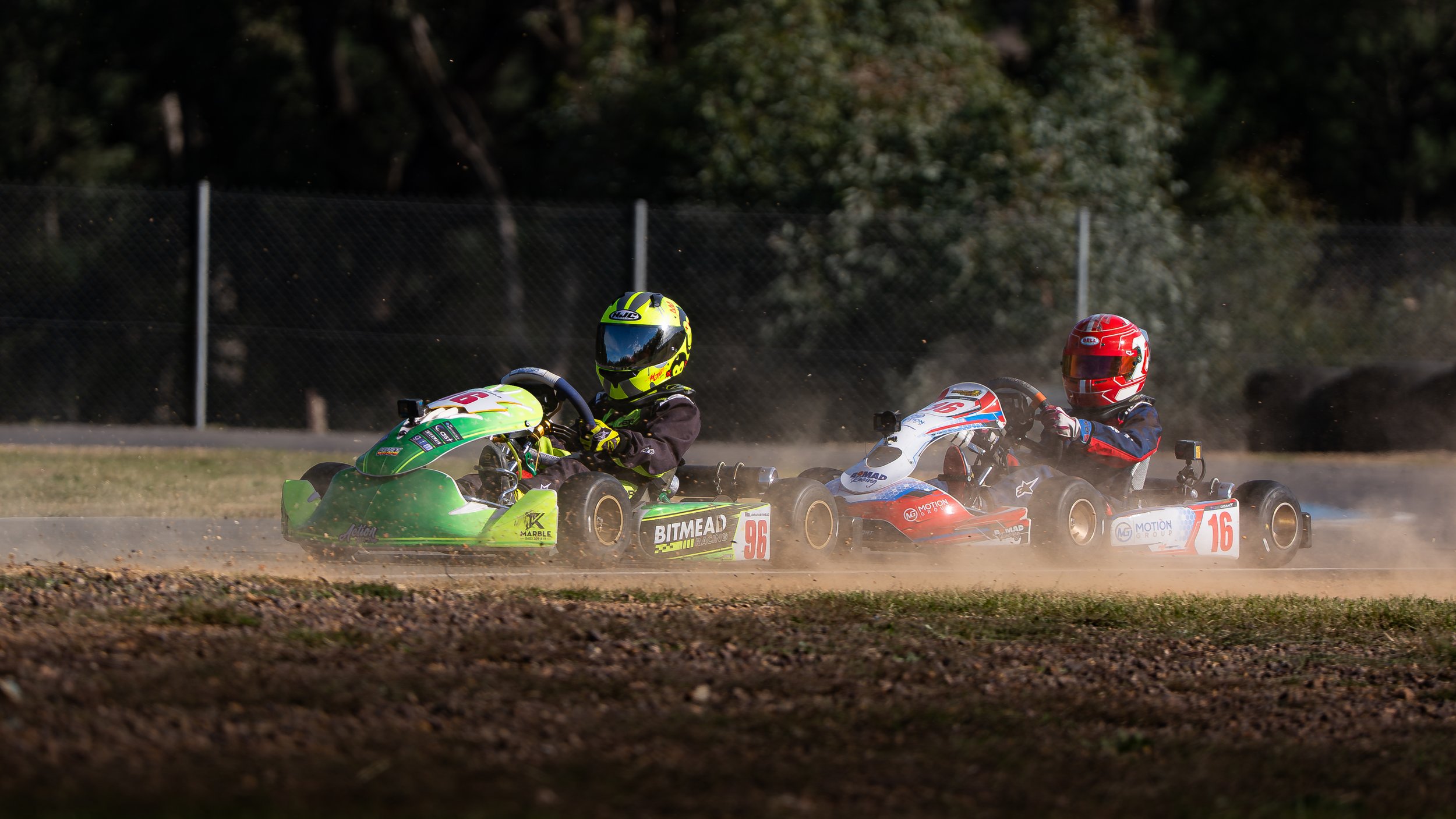 Two go-kart racers competing on a dirt track at dusk, both wearing helmets and race gear, kicking up dust behind them.