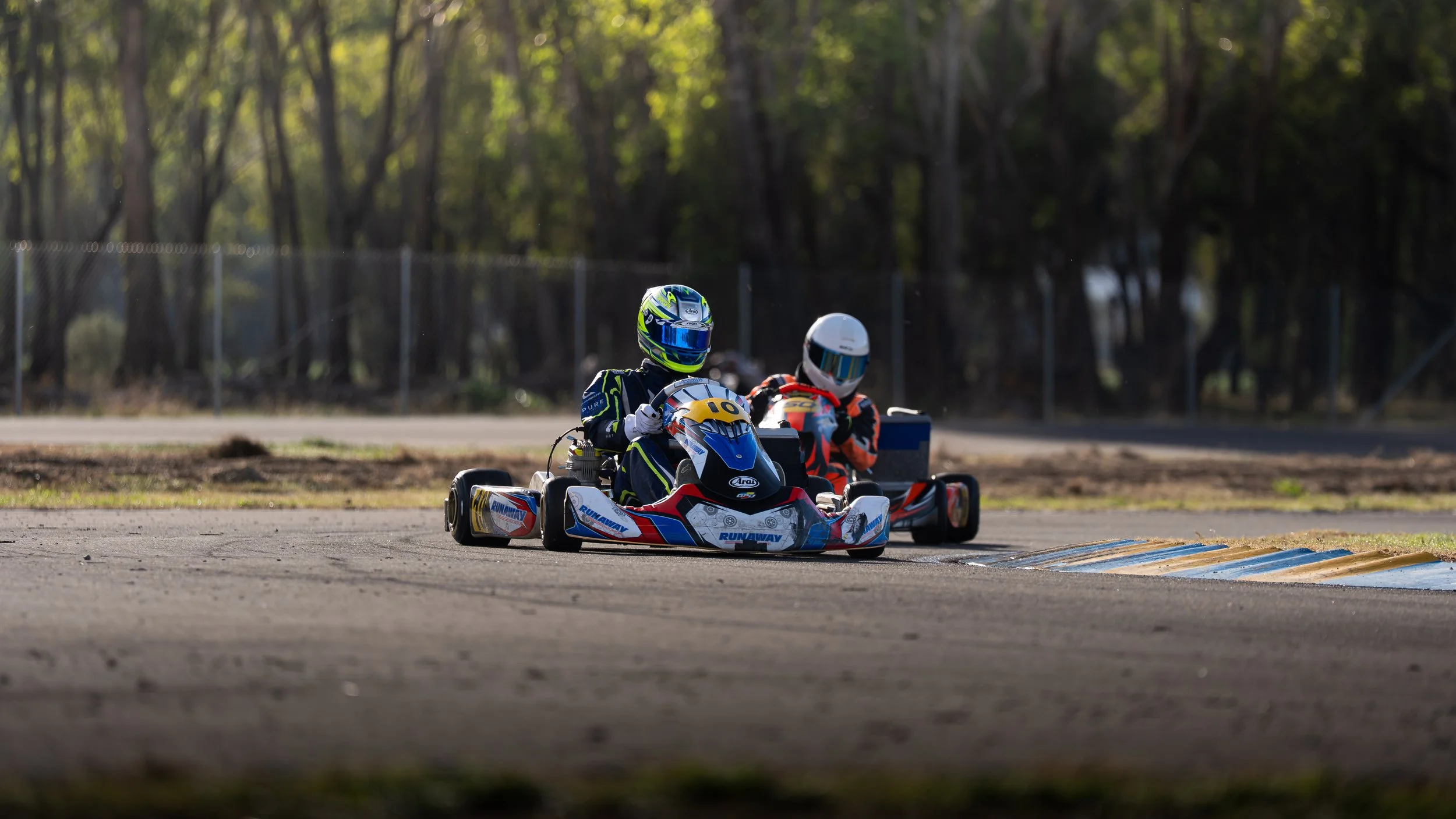 Two go-kart racers in racing suits and helmets sitting in their go-karts on a race track during daytime.