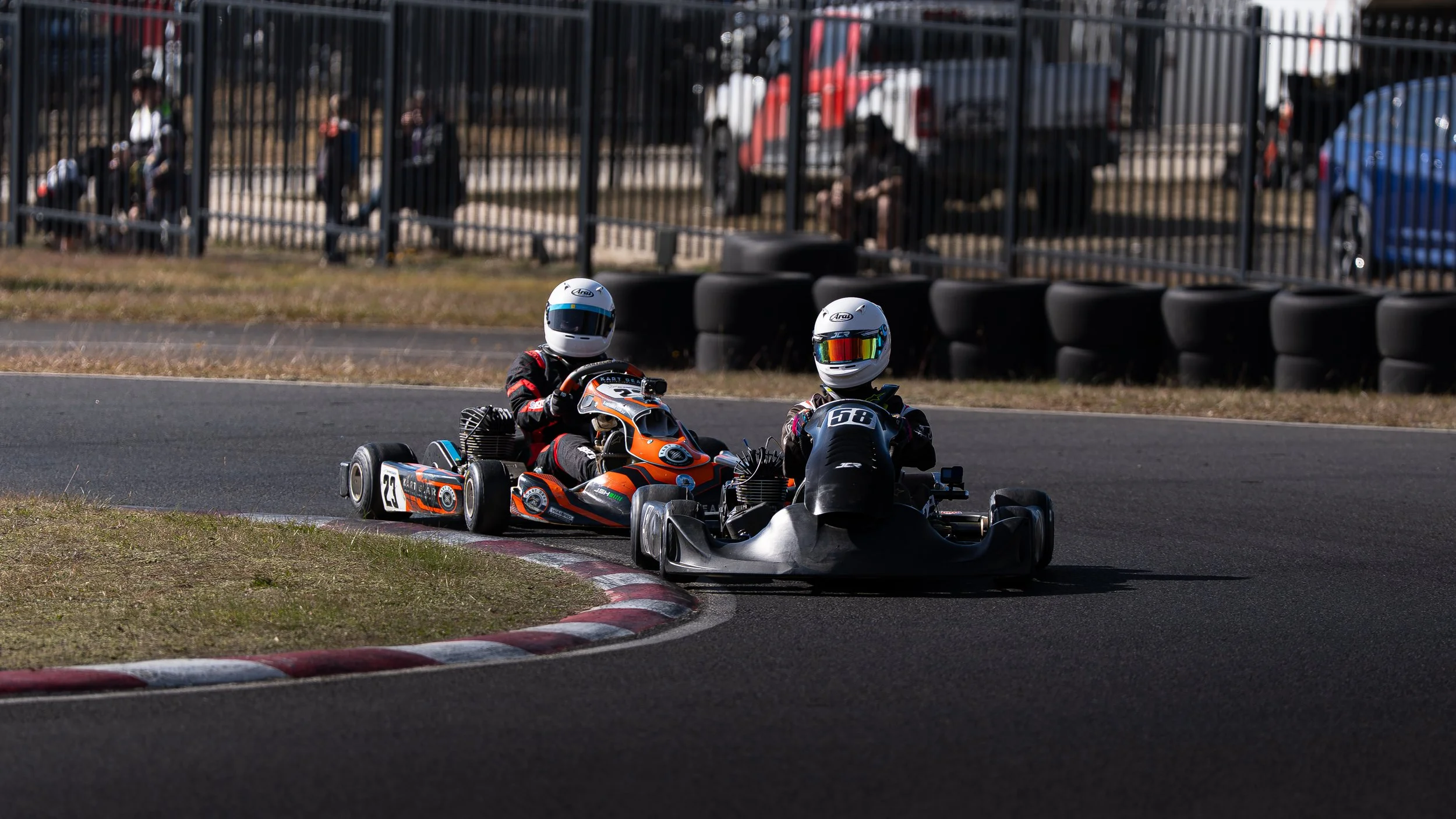 Two go-kart racers wearing helmets and racing suits on an outdoor track, with spectators behind a fence in the background.