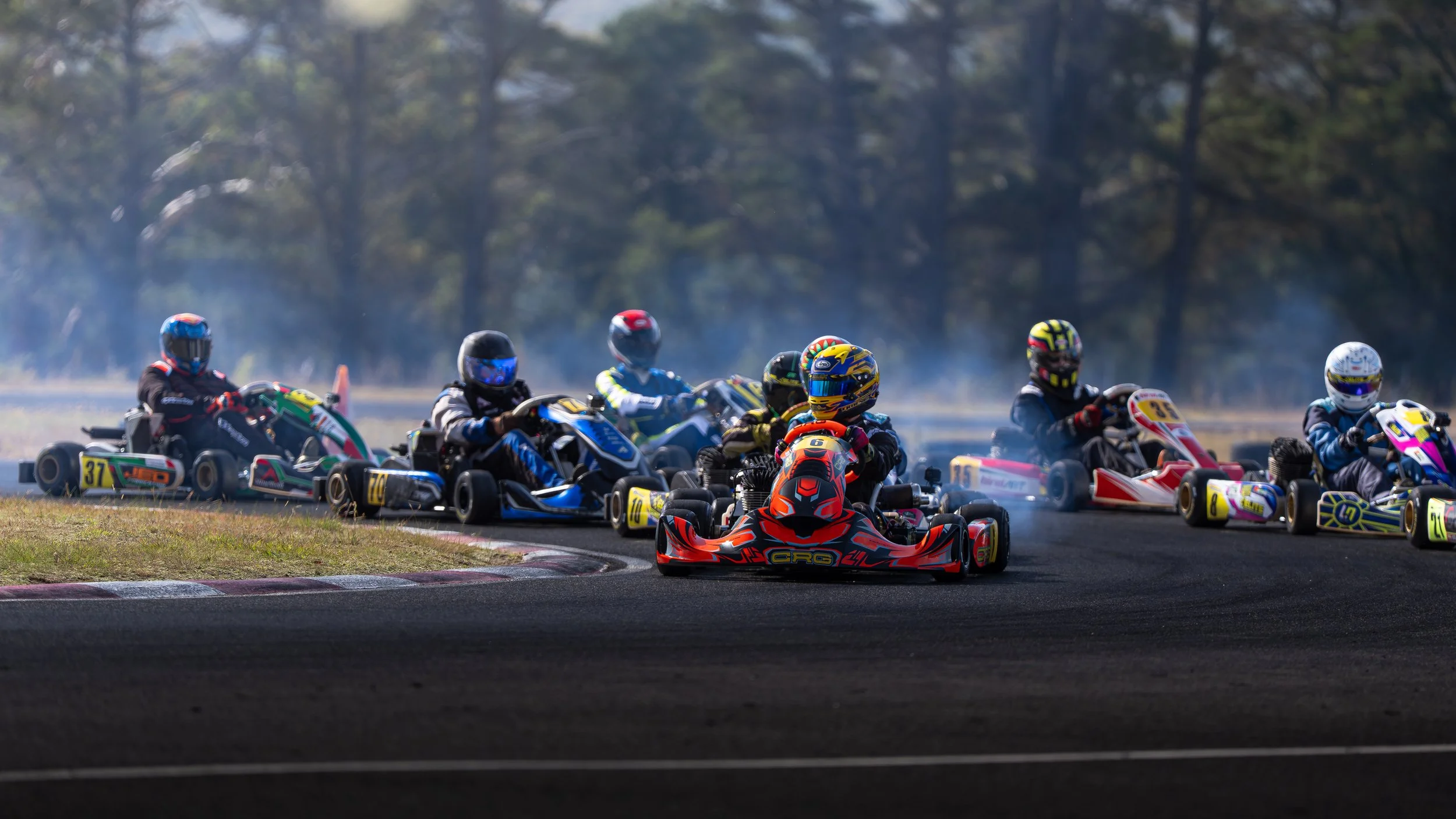 Several go-karts with drivers in racing helmets and suits navigate a turn on a race track, surrounded by a grassy area and trees in the background.