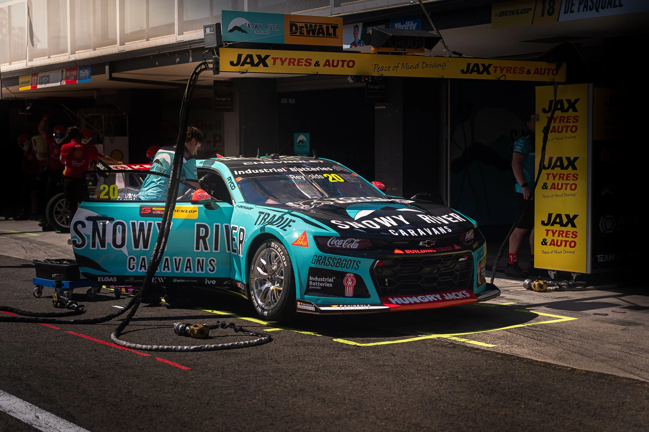 Race car parked in the pit lane at a racing event, with crew members working around it and various sponsor banners overhead.