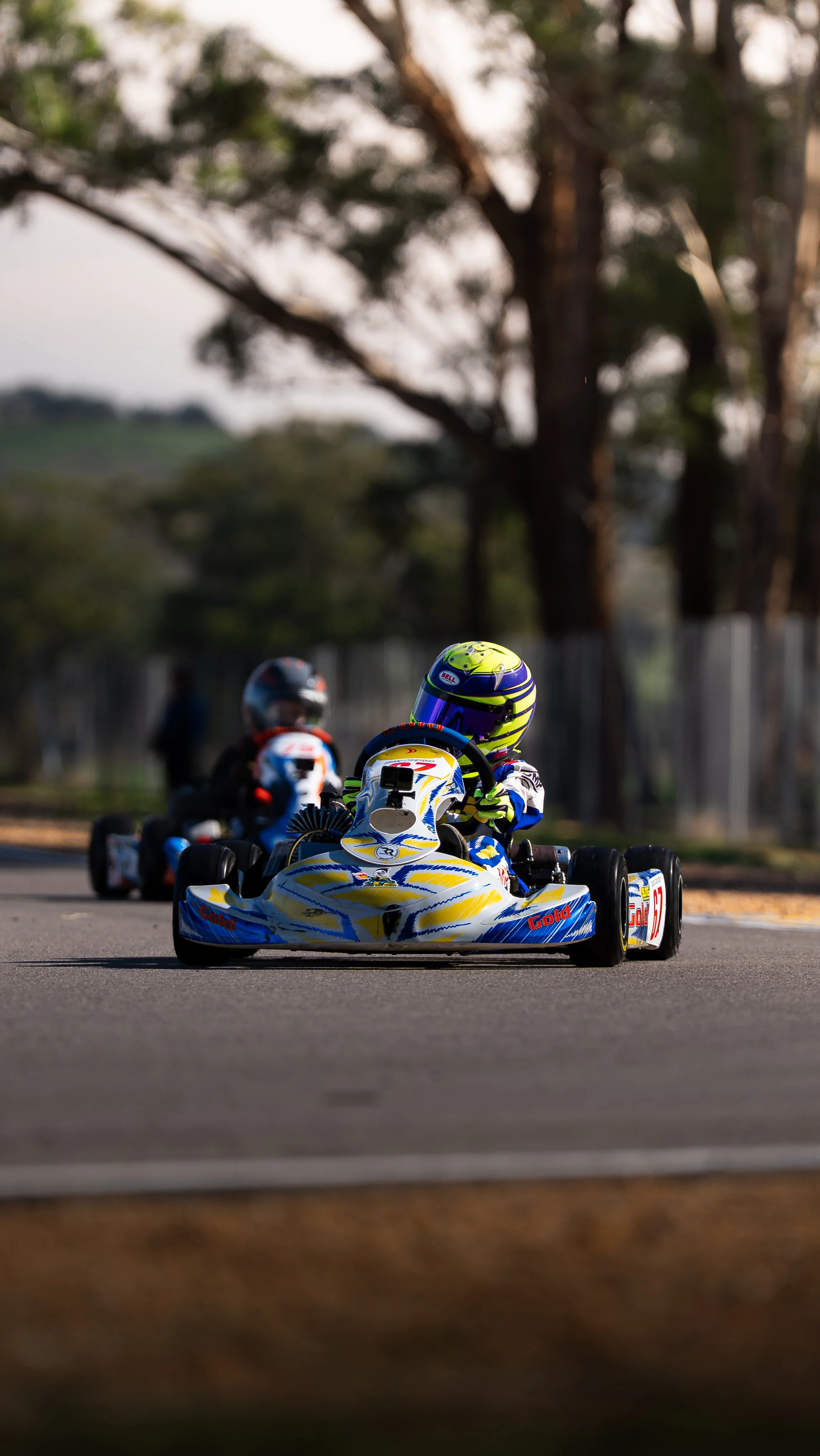 Go-kart racers on the track with trees and a fence in the background.
