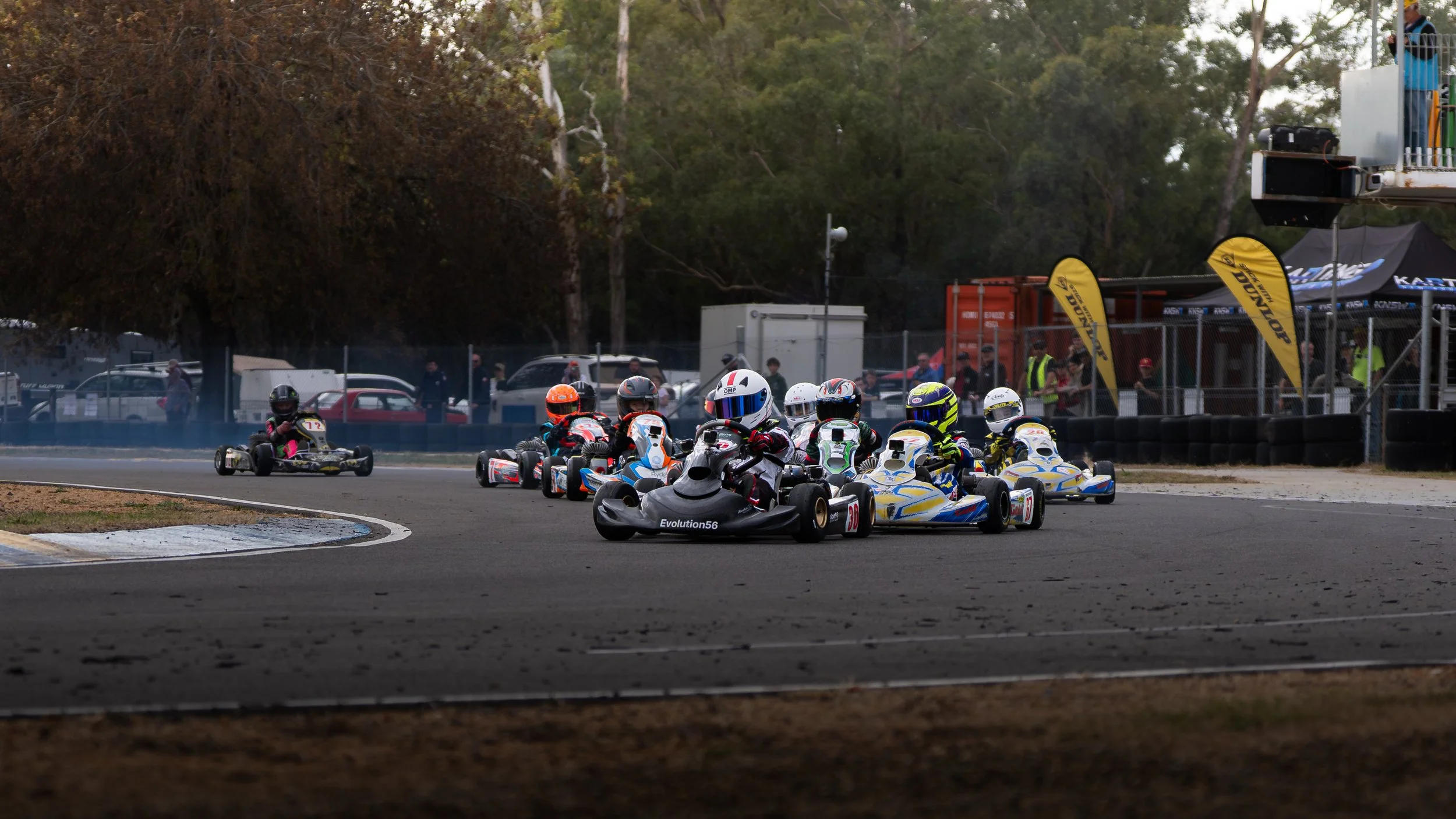 Go-kart race with multiple drivers navigating a turn on a racetrack, surrounded by trees, fences, and race banners.