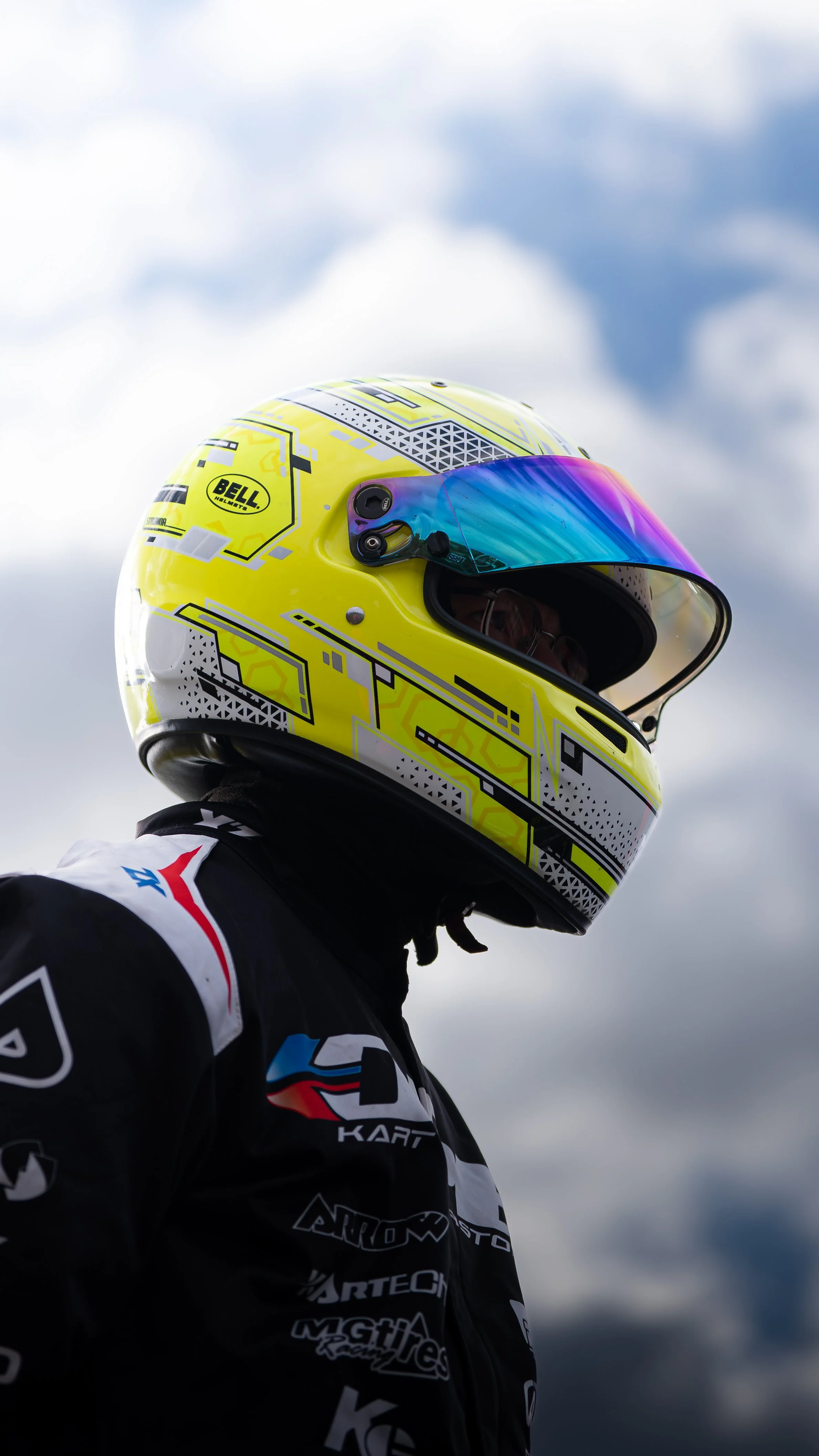 Close-up of a race car driver wearing a yellow racing helmet with a rainbow visor and a racing suit with various sponsor logos, standing outdoors against a cloudy sky.
