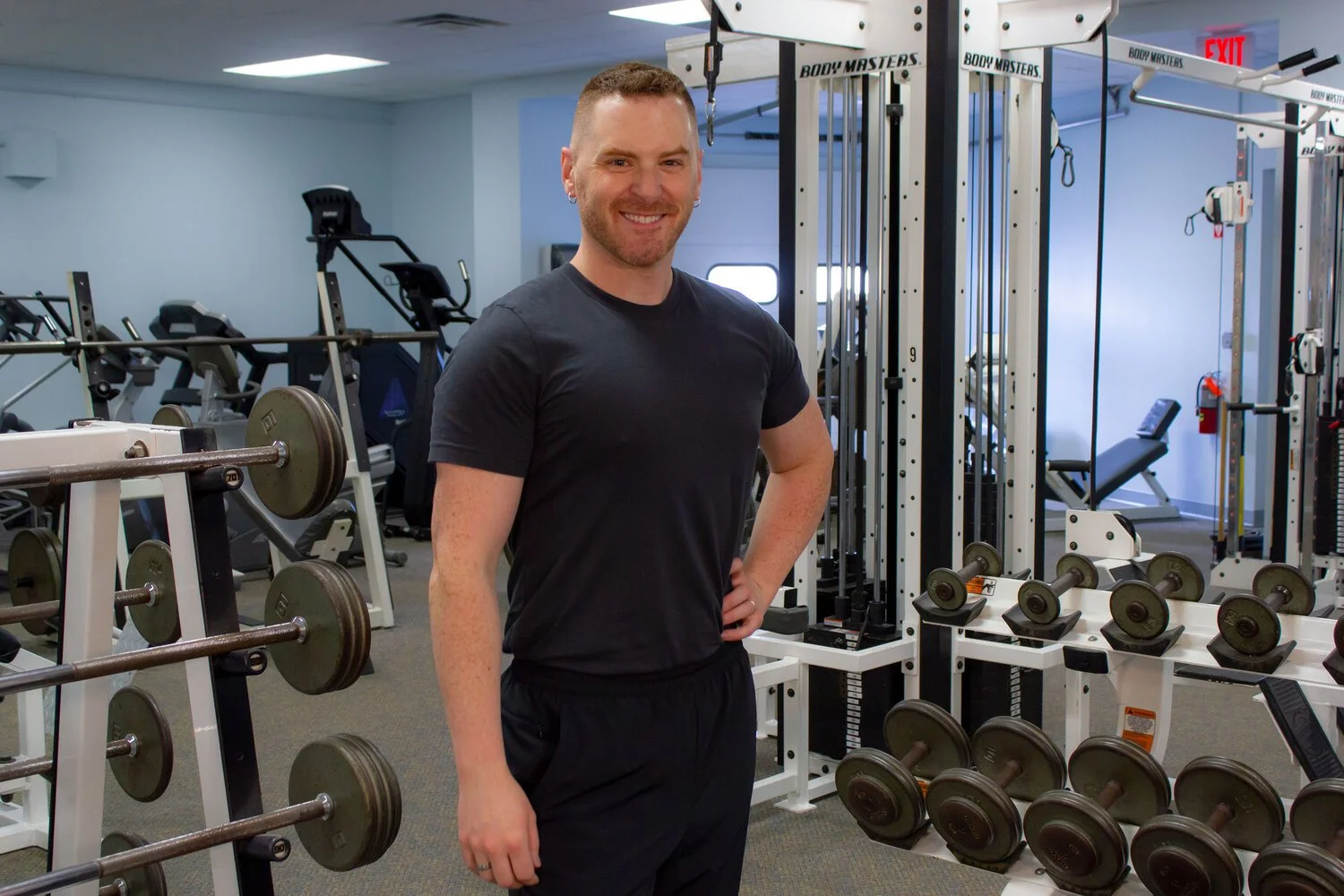 A smiling man standing in a gym, surrounded by various workout equipment including dumbbells, weight machines, and benches.