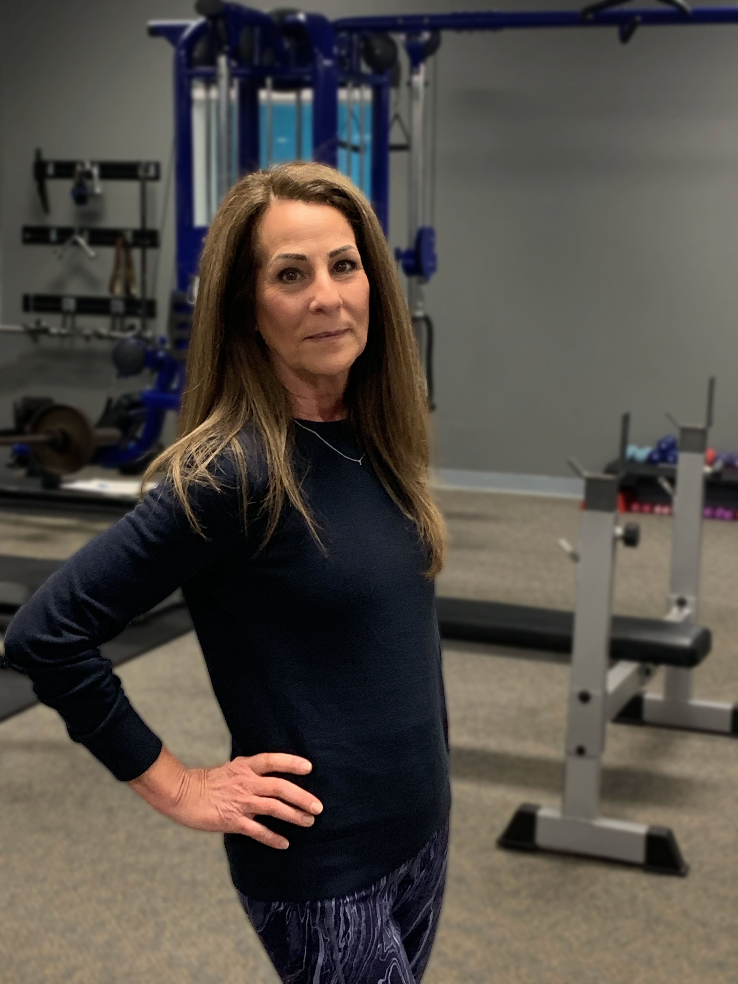 A woman with long brown hair standing in a gym with her hand on her hip, wearing a black long-sleeved shirt and patterned leggings.
