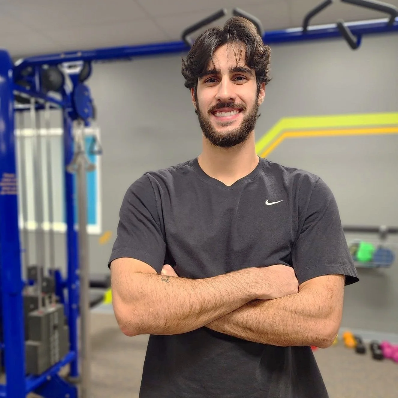 A young man with dark hair and a beard smiling, standing with arms crossed in a gym.