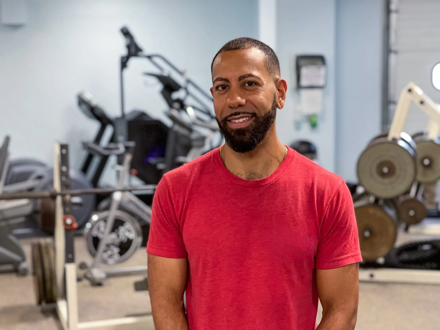 A man with a beard and short hair standing in a gym, smiling, wearing a red t-shirt.