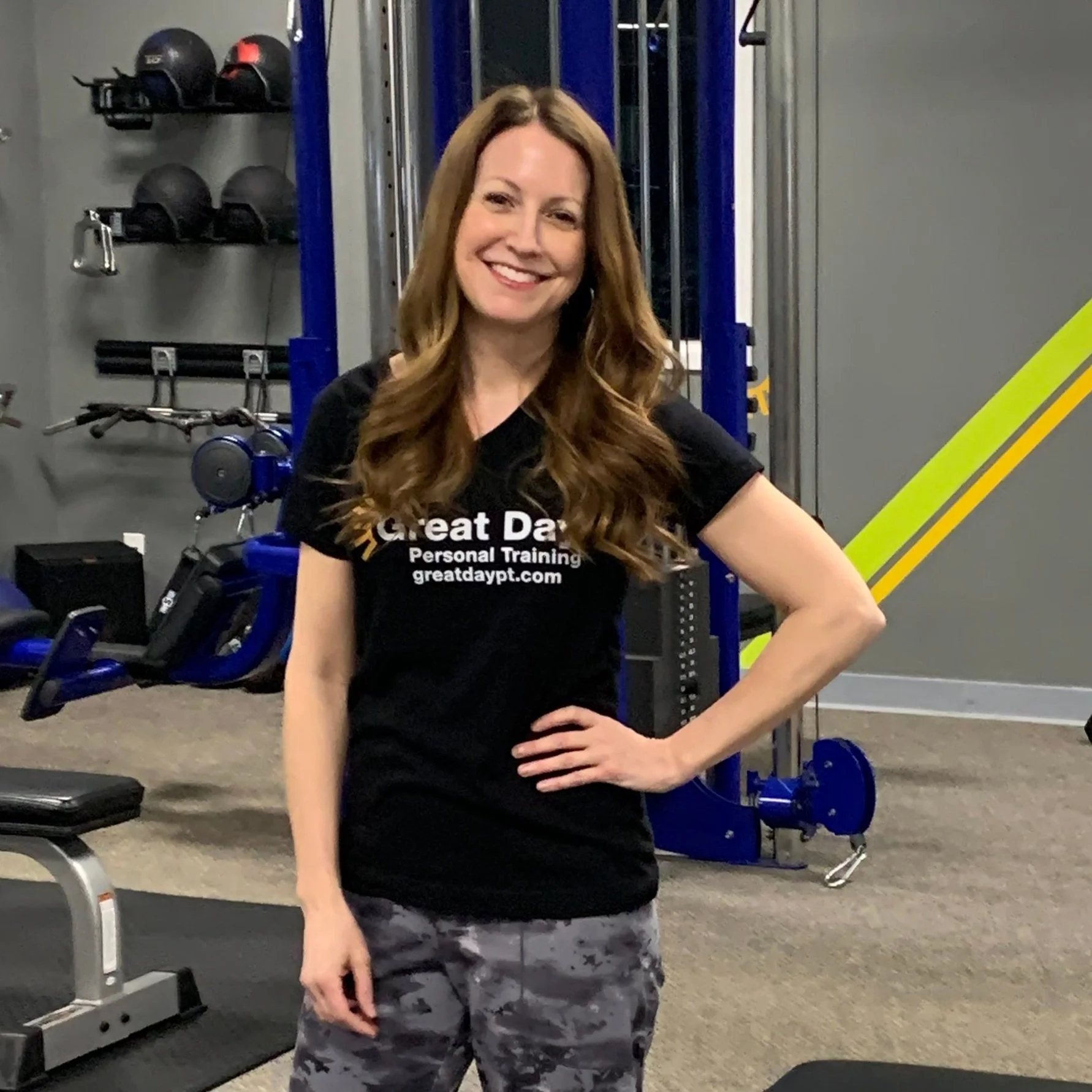 A woman with long, wavy red hair smiling and standing confidently in a gym, wearing a black T-shirt and camouflage-patterned workout pants.