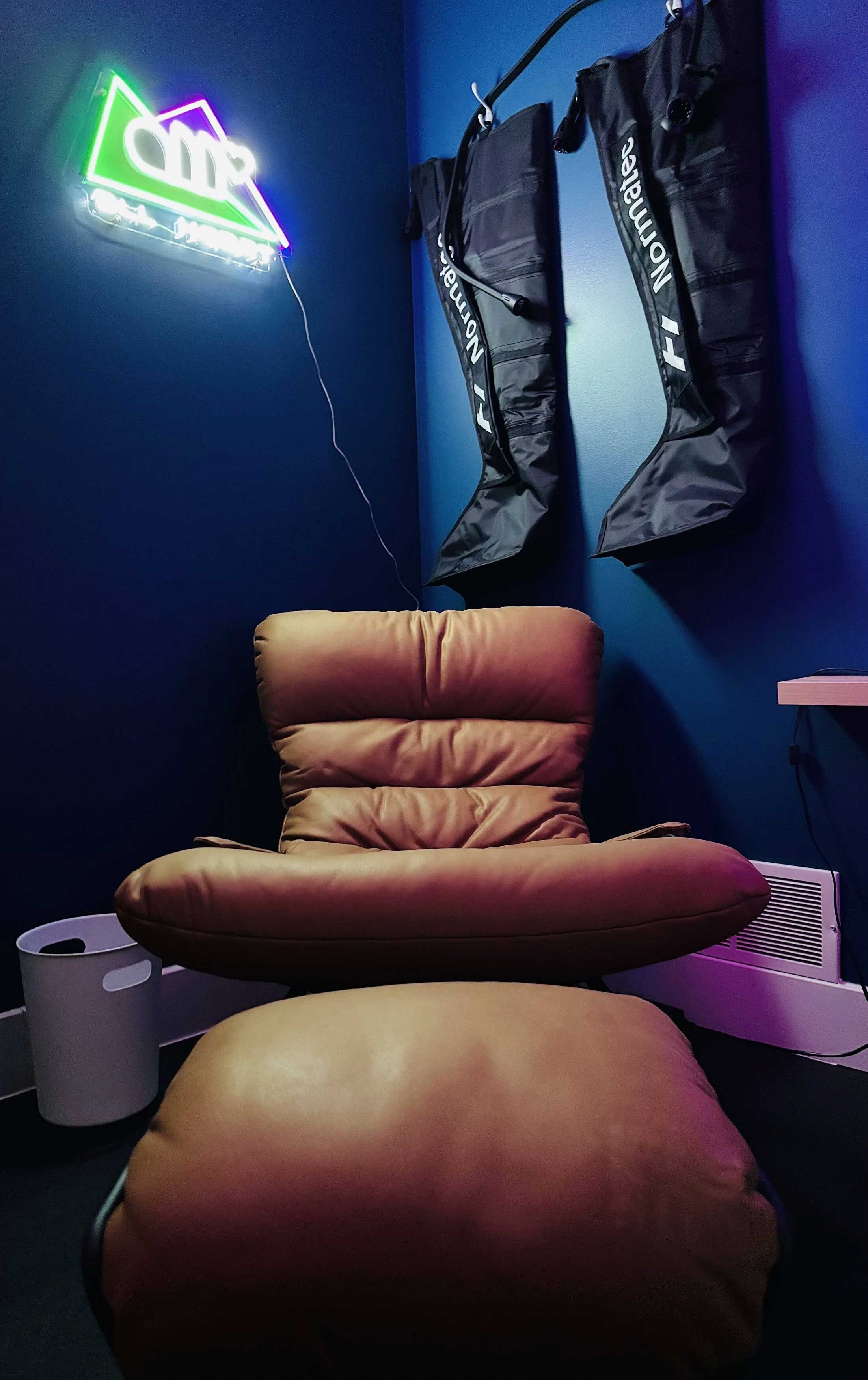 A relaxation corner with a brown cushioned lounge chair and matching ottoman in a room with dark blue walls. Two black NoMoreGear hanging storage bags are mounted on the wall, and a neon sign with colorful lights is visible, illuminating part of the 