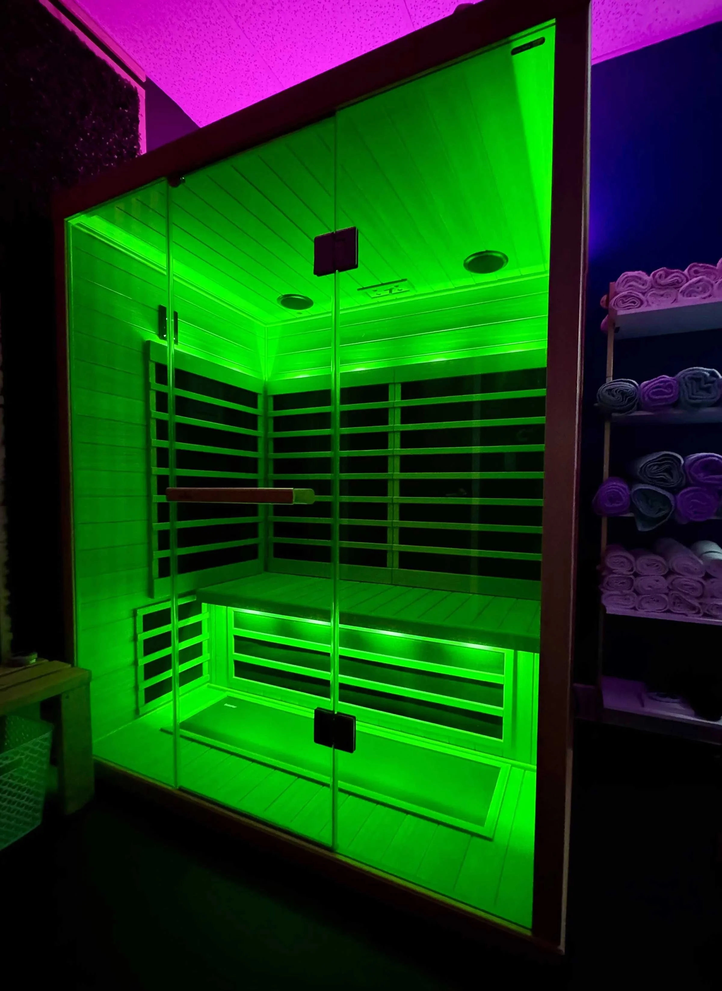 An infrared sauna with green lighting, glass doors, and wooden interior, situated next to a shelf with rolled towels.