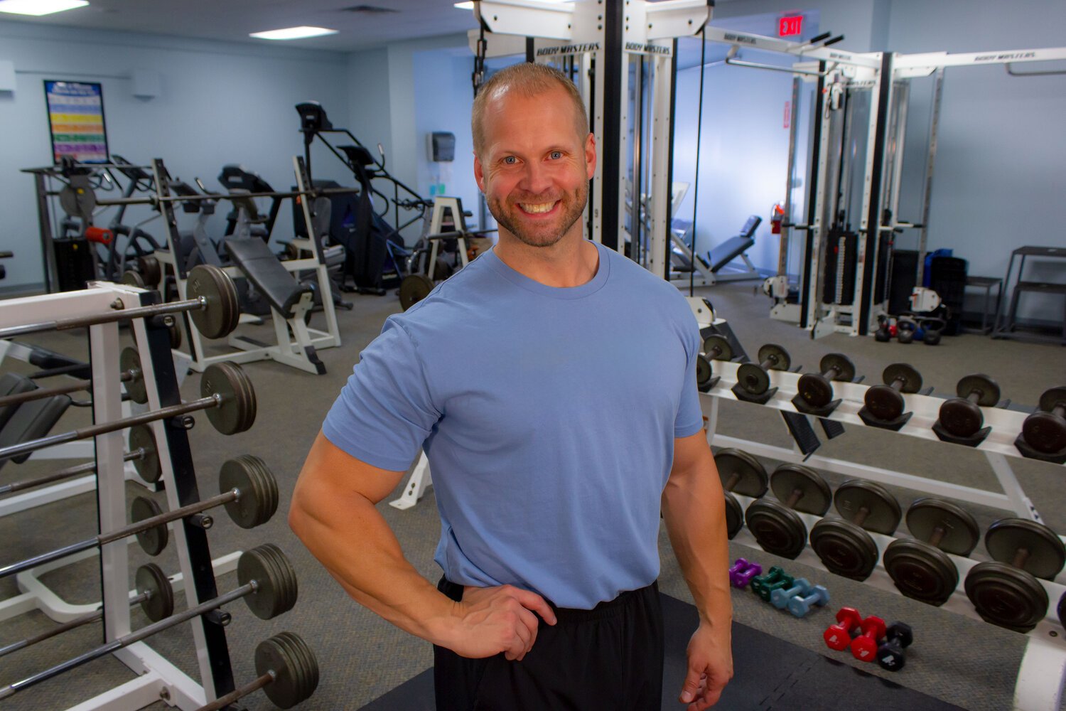 A smiling man in a light blue t-shirt standing in a gym with various exercise equipment, including dumbbells and weightlifting machines, in the background.