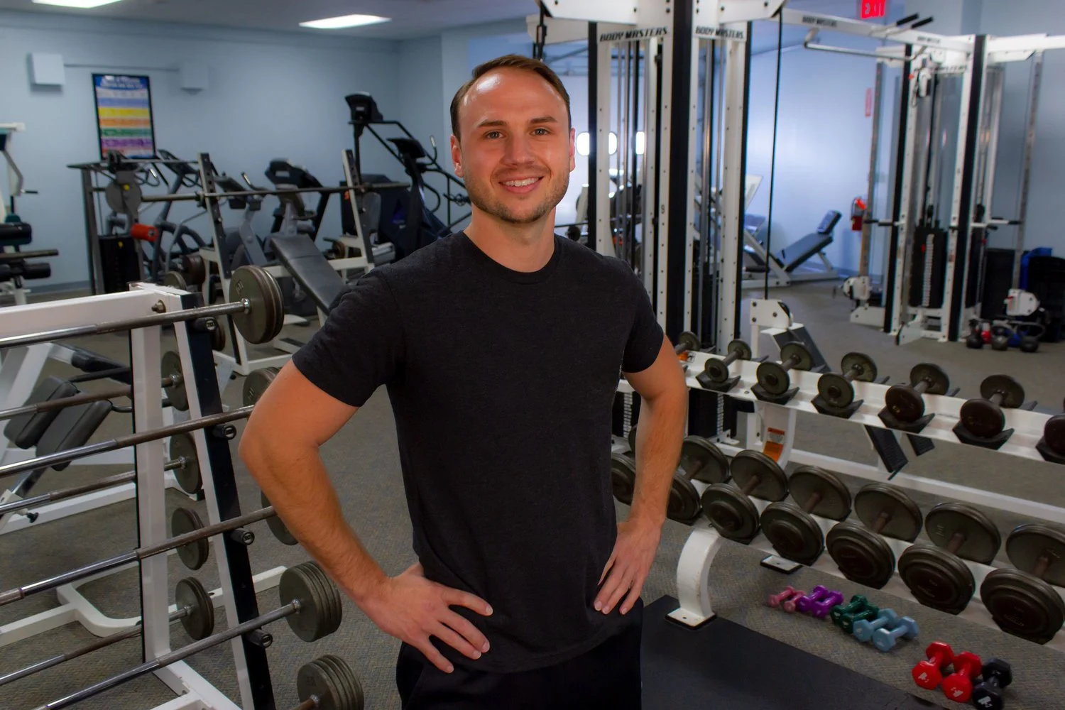 A smiling man in a black t-shirt standing with hands on hips in a gym, surrounded by weightlifting equipment and dumbbells.