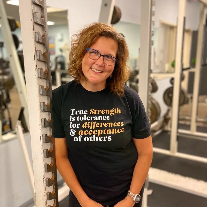 A woman with curly red hair and glasses smiling in a gym, standing next to weightlifting equipment, wearing a black T-shirt with motivational words.