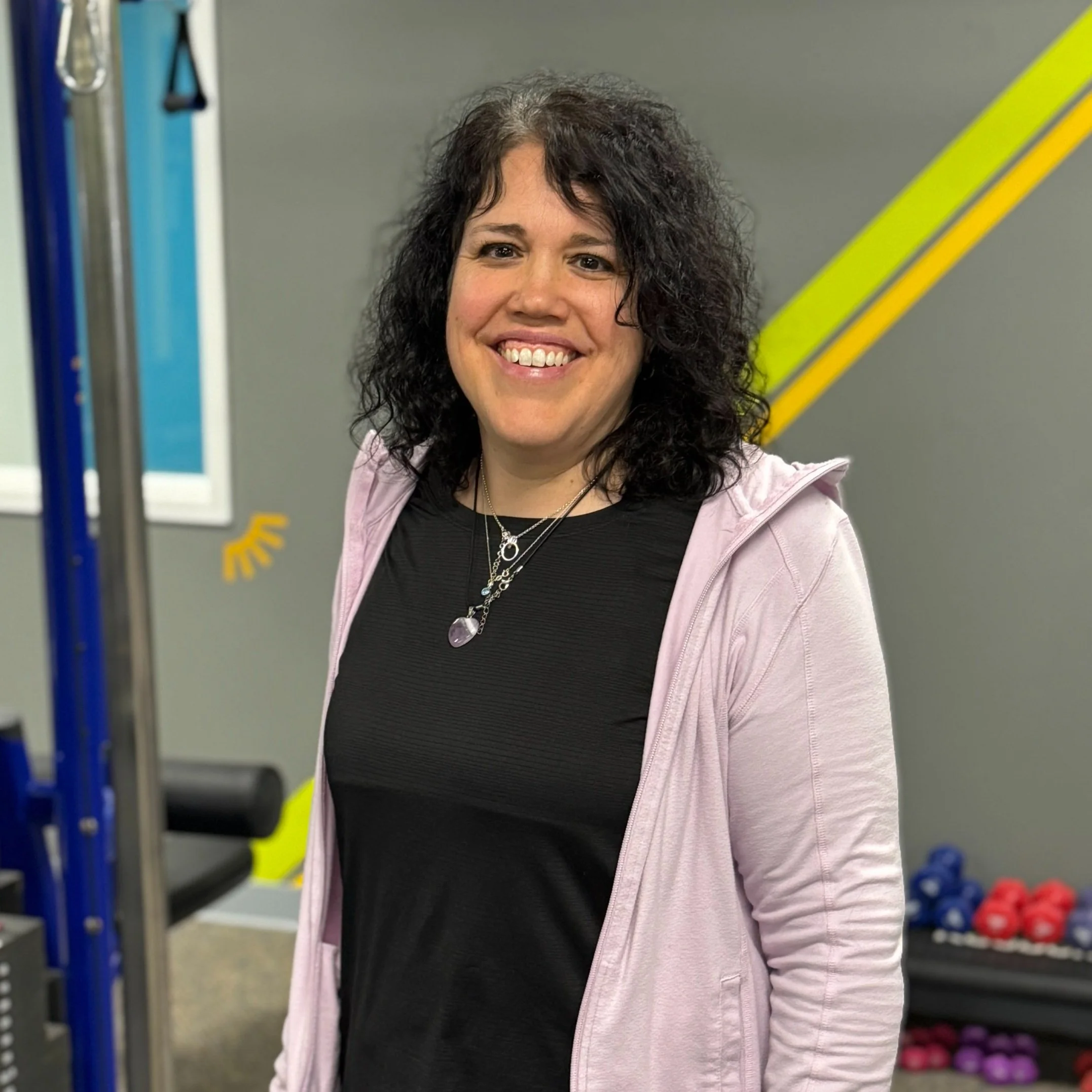 Smiling woman with curly dark hair wearing a black shirt, pink jacket, and layered necklaces in a gym with workout equipment in the background.