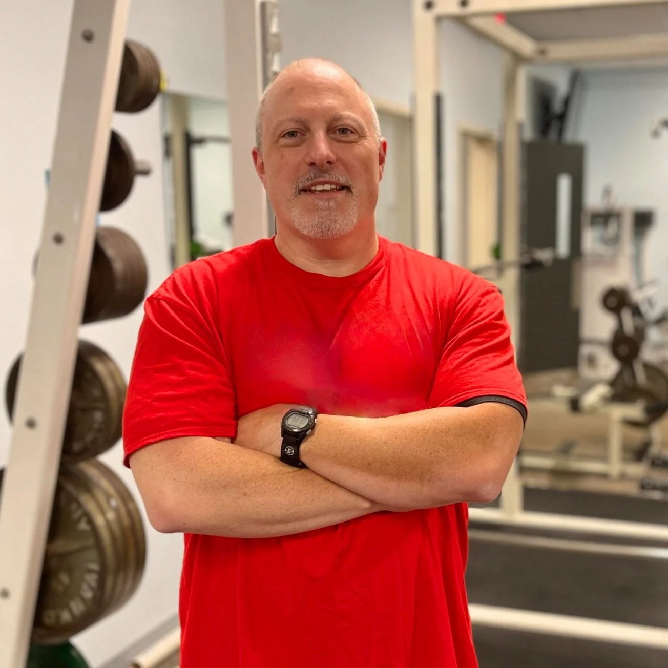 A middle-aged man with a bald head, beard, and mustache wearing a red T-shirt and a black digital watch, standing with arms crossed in a gym with weight racks and workout equipment in the background.