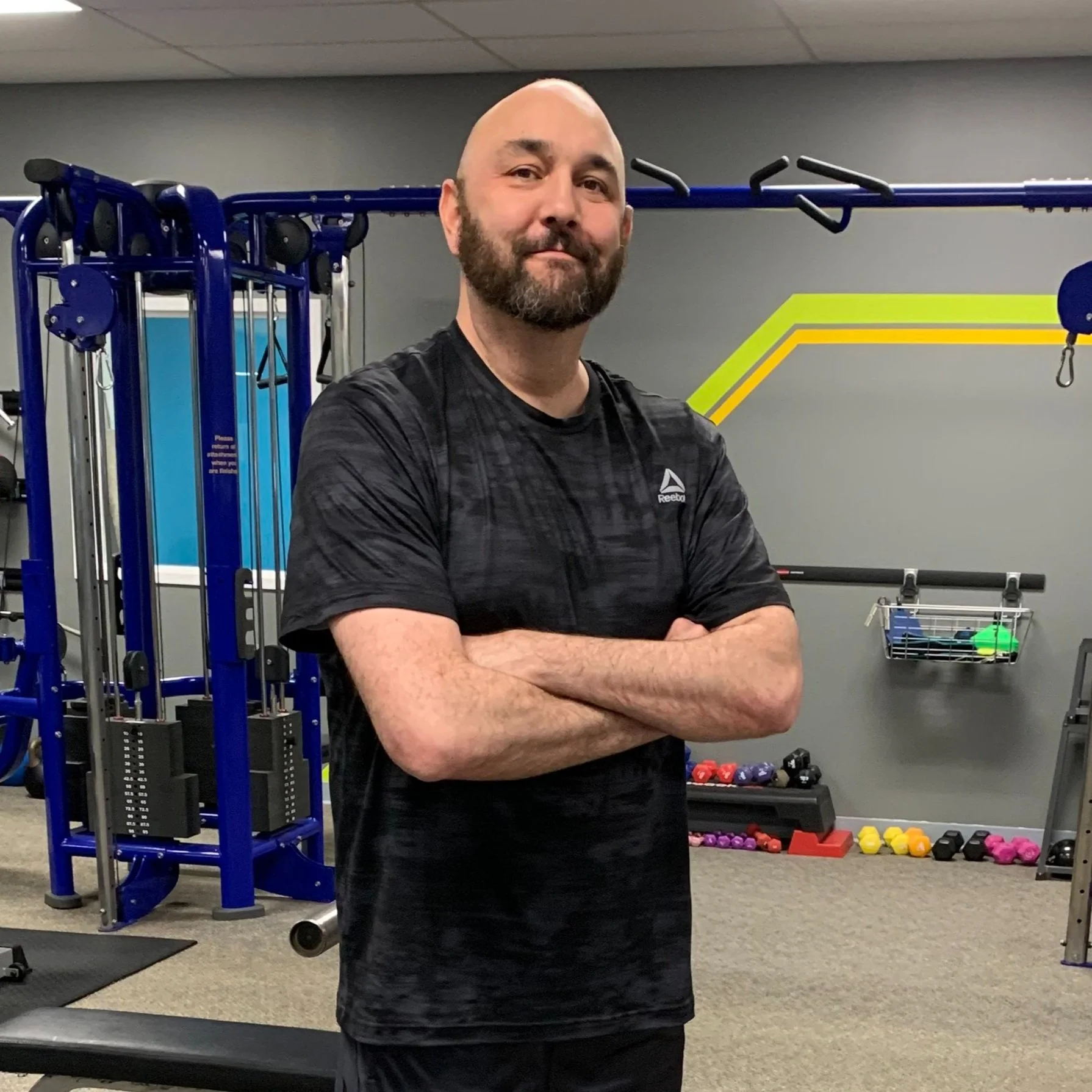 A man with a beard and bald head standing with arms crossed in a gym, smiling slightly. Gym equipment like a blue multi-station workout machine, dumbbells, and colorful workout gear are visible in the background.