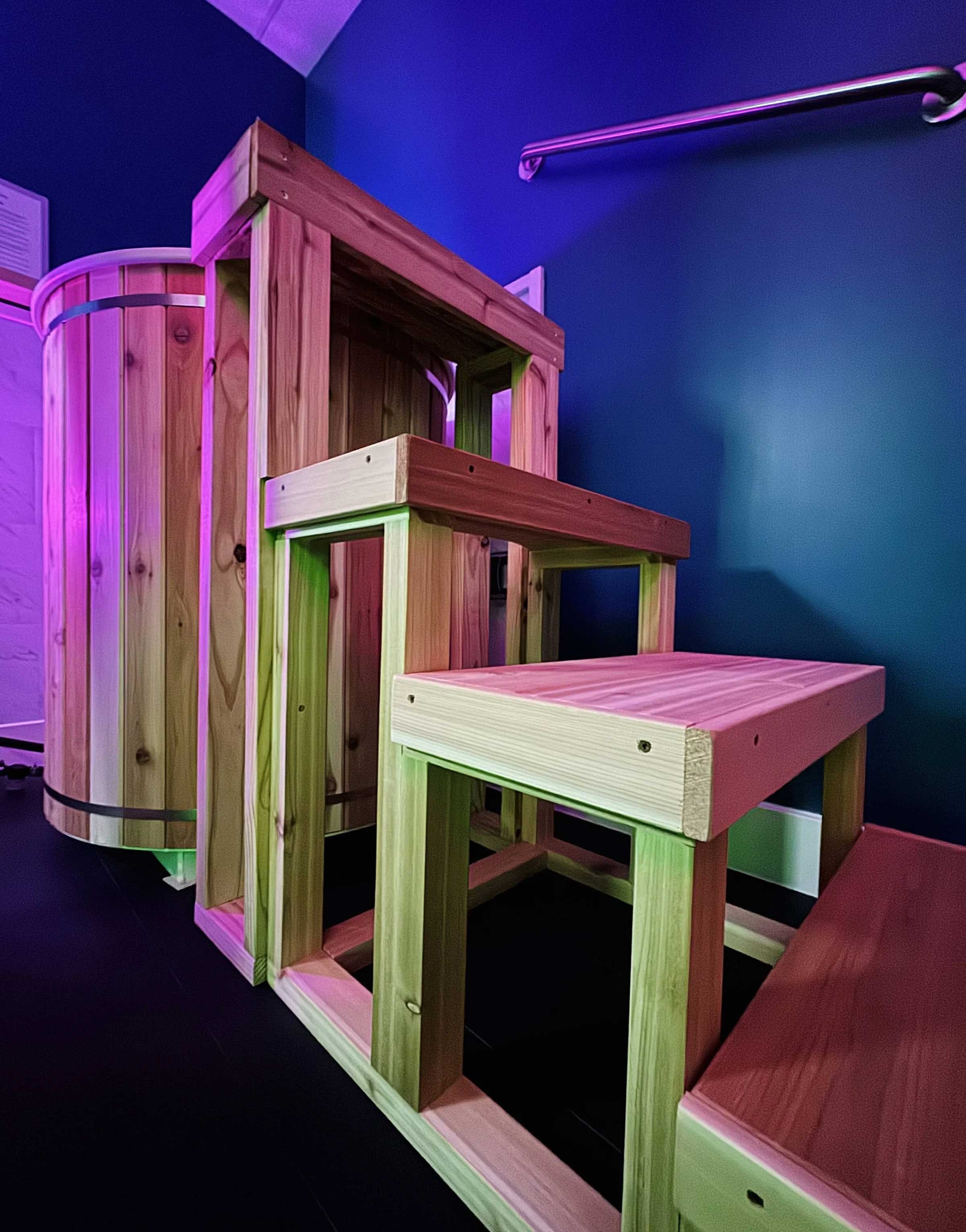 Indoor play structure made of unfinished wood, with stairs and a small platform, in a room with dark blue walls and purple lighting.