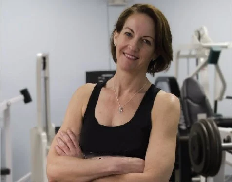 A woman with short brown hair smiling, standing in a fitness gym with workout equipment in the background.