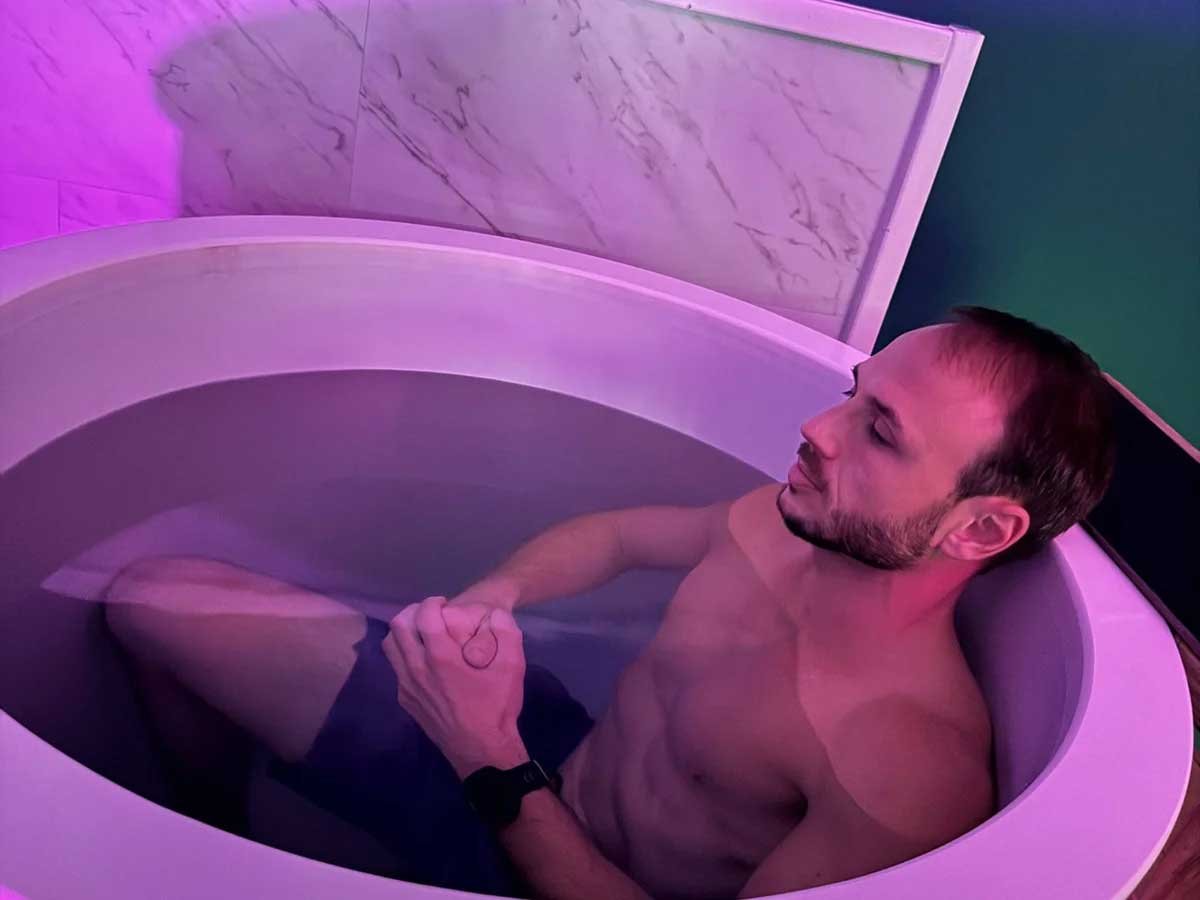A shirtless man with a beard relaxing in a purple-lit hydrotherapy bathtub.
