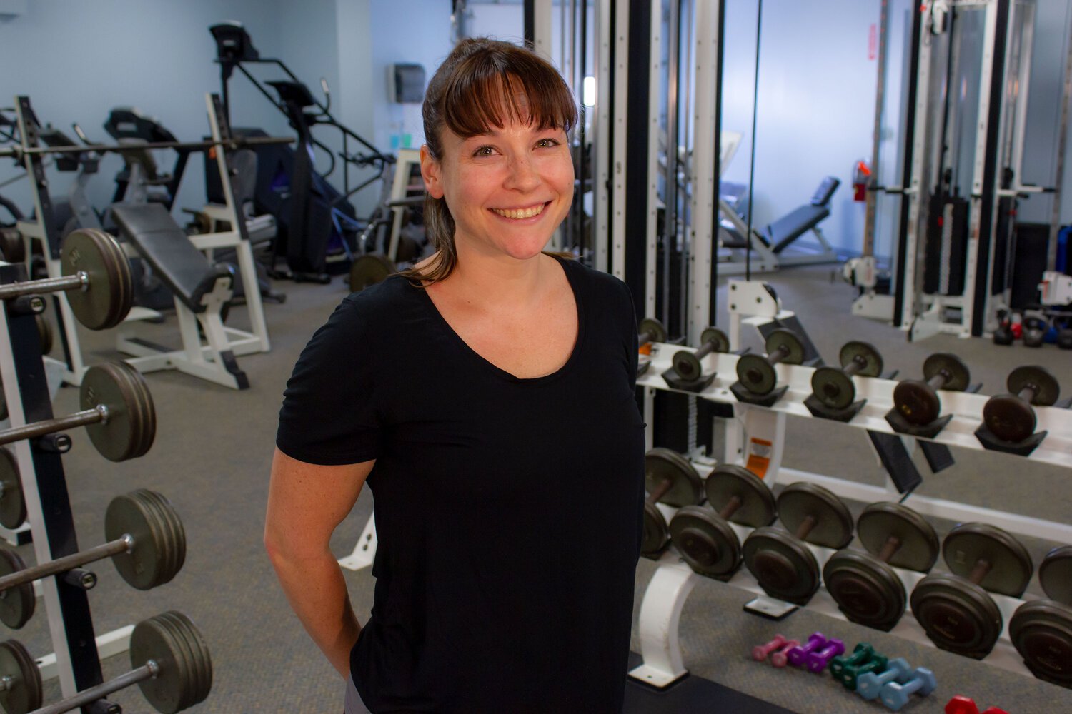 A woman smiling at the camera in a gym, standing near a rack of dumbbells and fitness equipment.