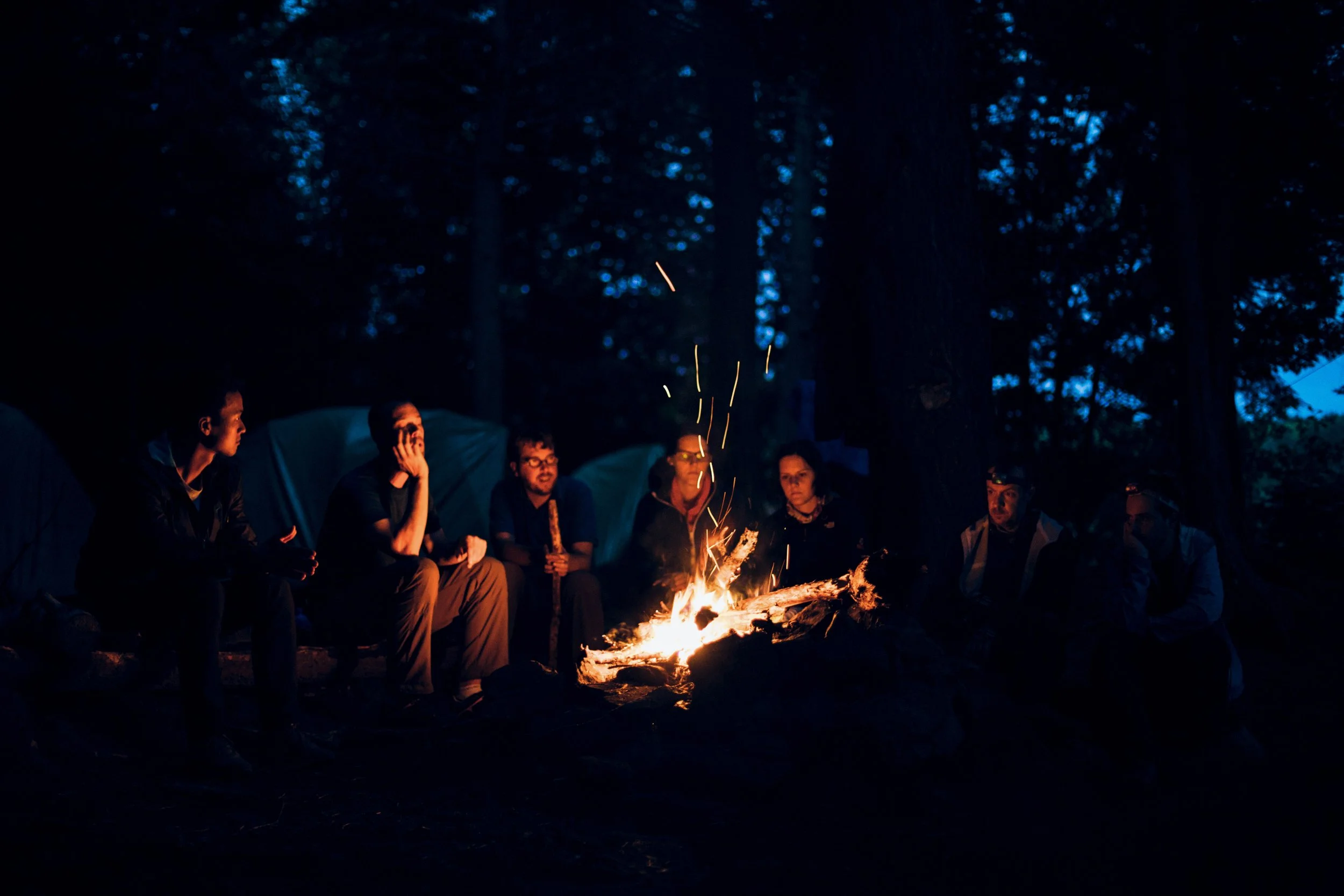Group of people sitting around a campfire in the Adirondacks enjoying a close up magic show at their campsite.  The magician captivates the group in an extremely comfortable and exclusive setting for magic.