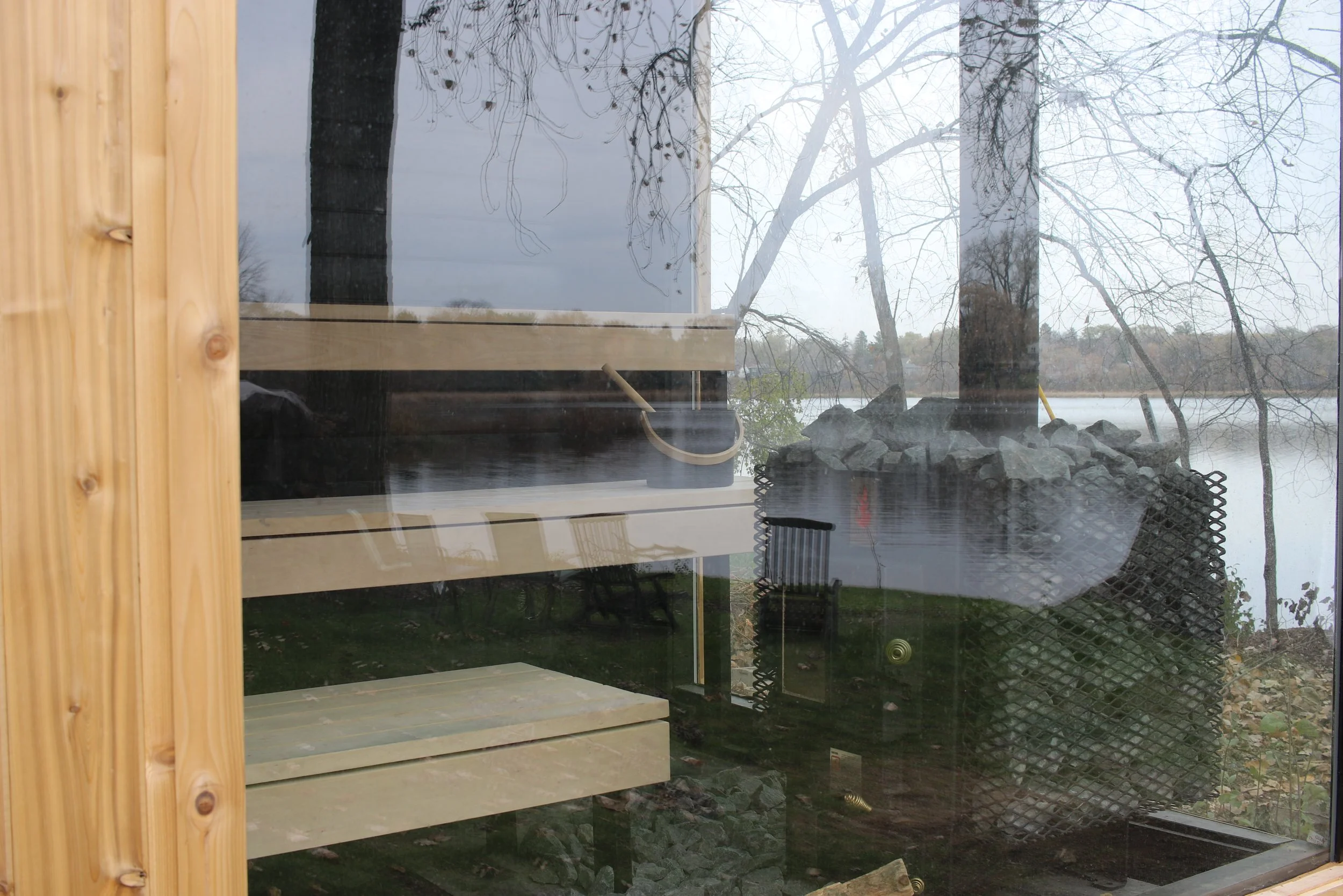 View through a large glass window showing a lake, leafless trees, and a cloudy sky, with reflections of outdoor chairs and landscaping.