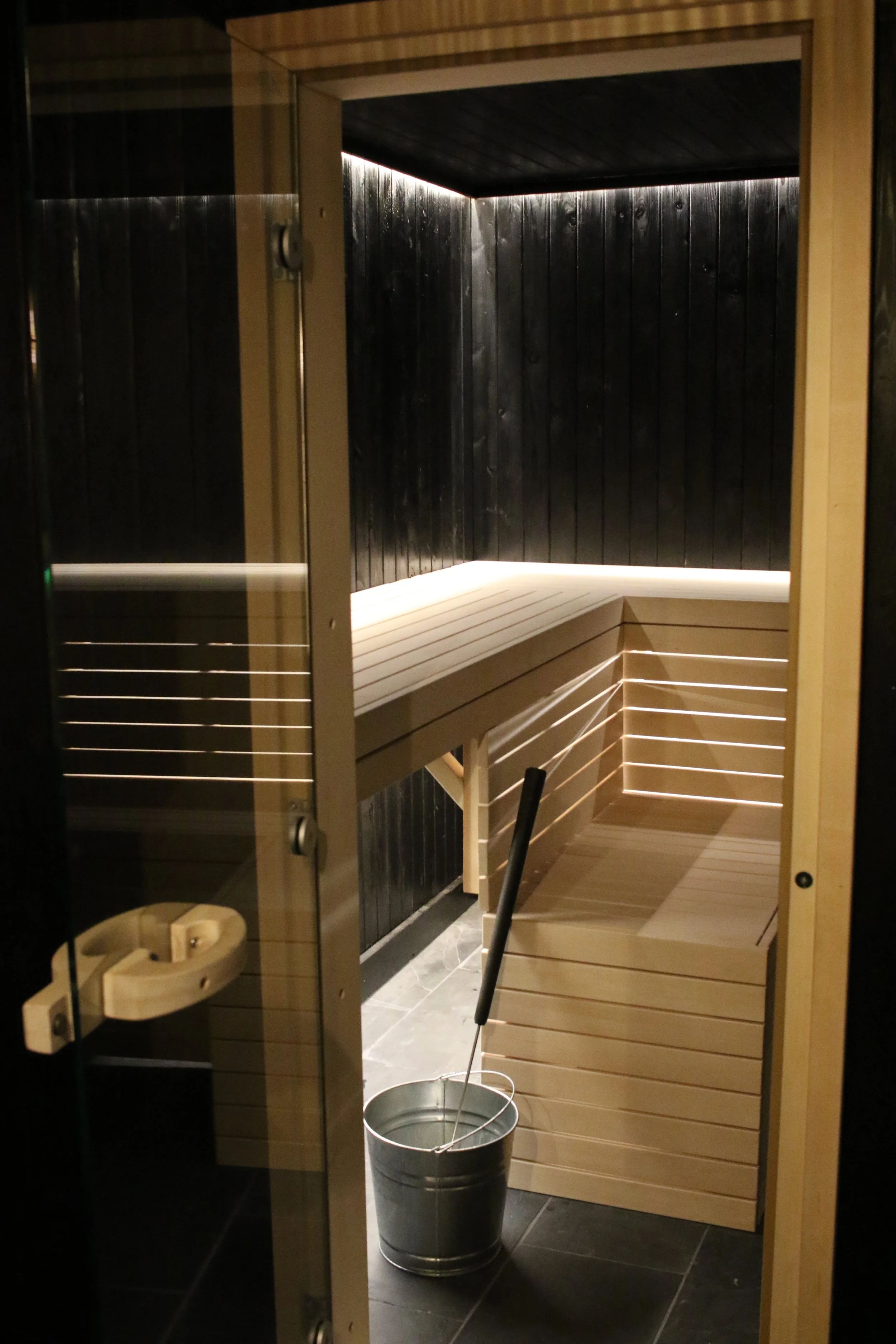 Modern infrared sauna with wooden benches and black paneled walls, bucket and mop on the floor.
