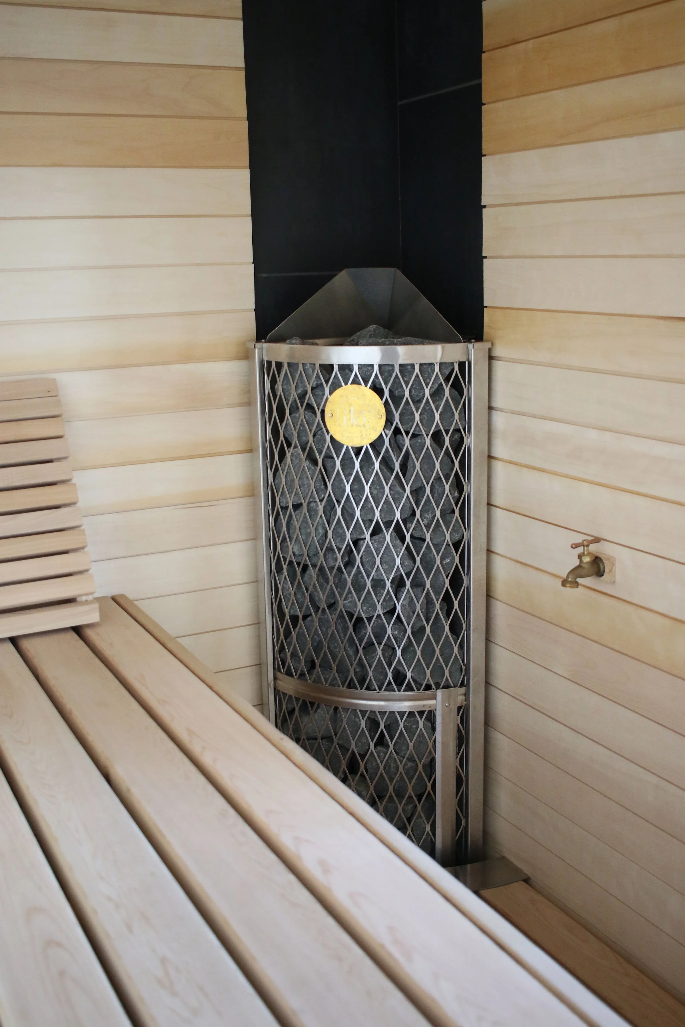Wooden interior of a sauna with a metal stove filled with rocks, a small water tap on the wall, and wooden benches.