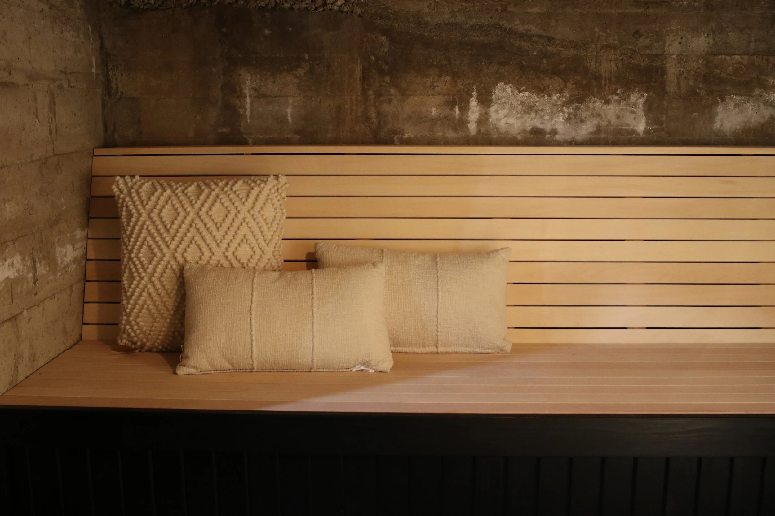 Wooden bench with three beige cushions against a slatted wooden backrest, in a room with concrete walls.