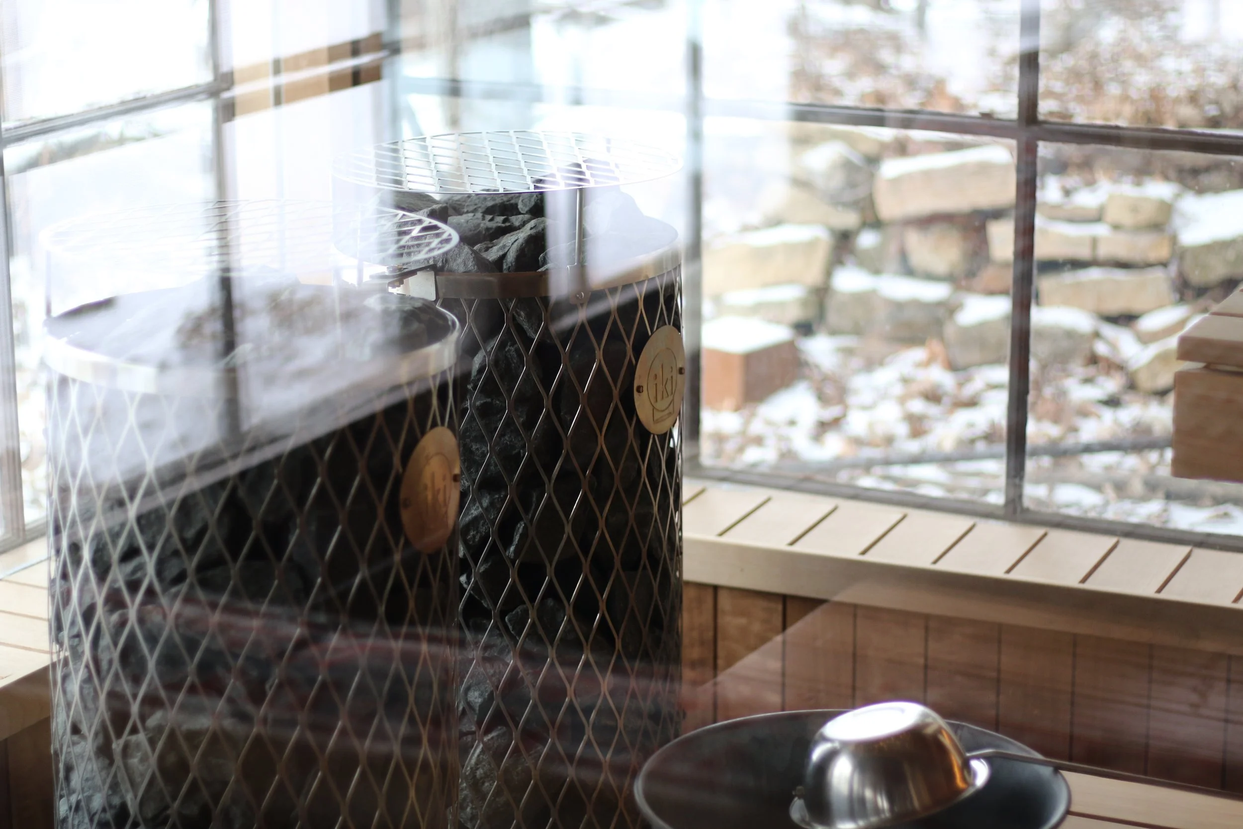 Inside a cozy structure, a wood stove with neatly stacked firewood behind a wire mesh, a metal bowl on a wooden surface, and a window showing a snowy outdoor scene.