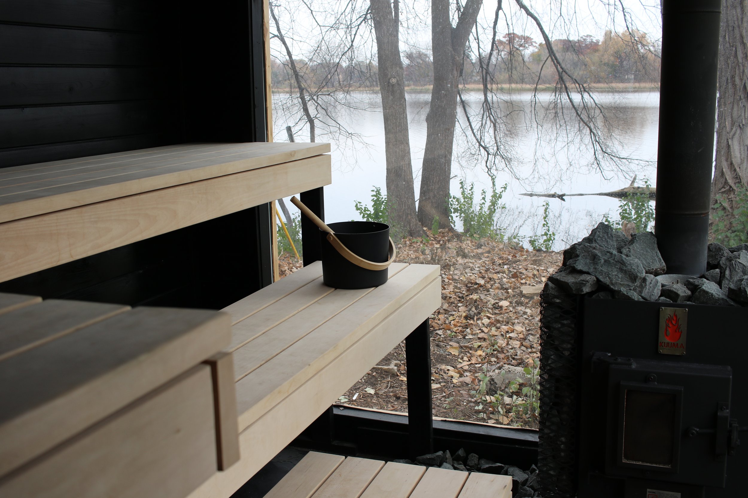 Interior of a sauna with wooden benches, a black bucket, and a view of a lake and leafless trees outside through a large window.