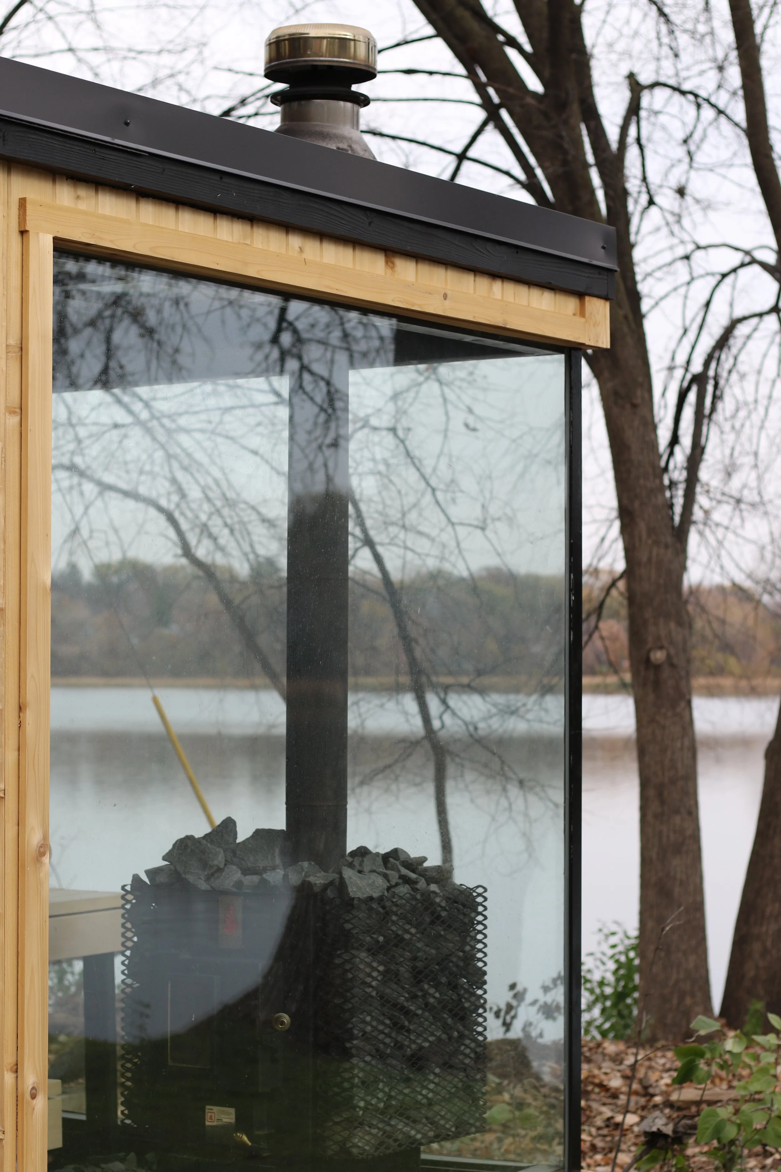 A small outdoor sauna or steam room with a glass front, located by a lake with trees and fallen leaves in the background.