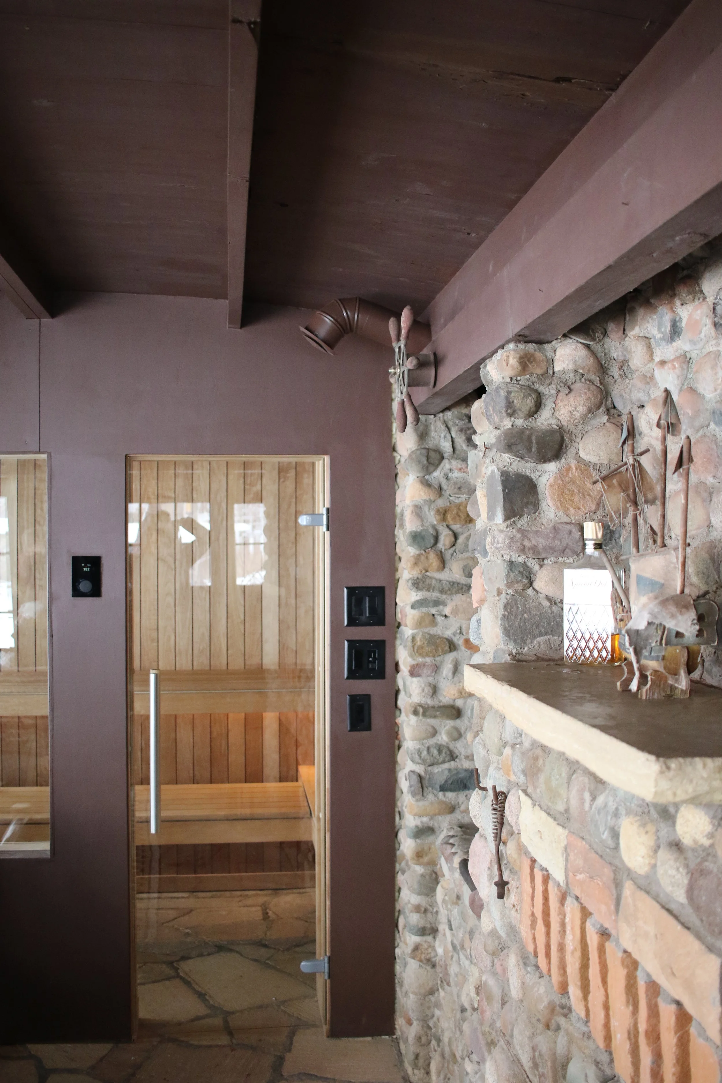Interior of a rustic sauna room with a stone wall, wooden door, and sauna interior visible through glass, with decorative metal tools on the wall.