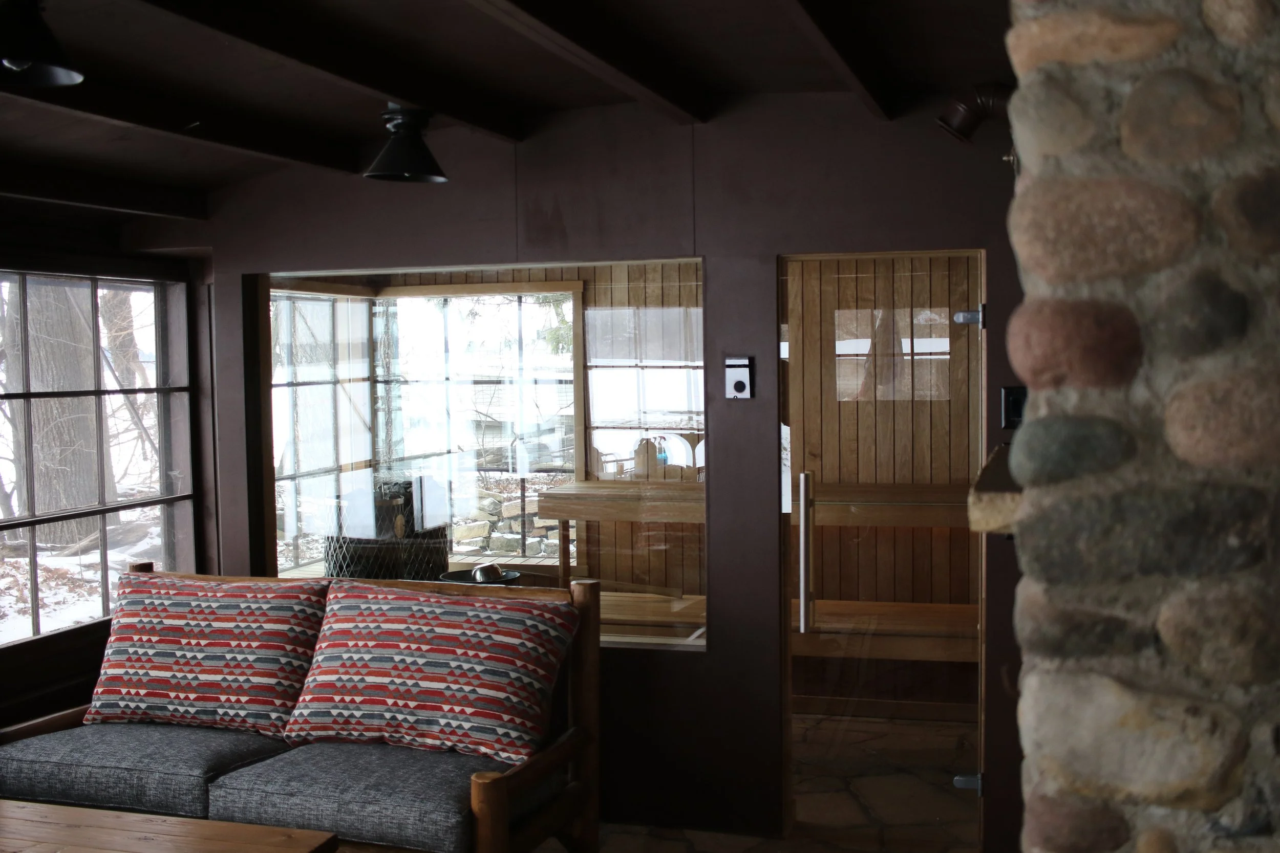 Interior of a cozy room with a stone wall on the right, a window with snow outside, a gray sofa with patterned pillows, and a wooden sauna room with a glass door.