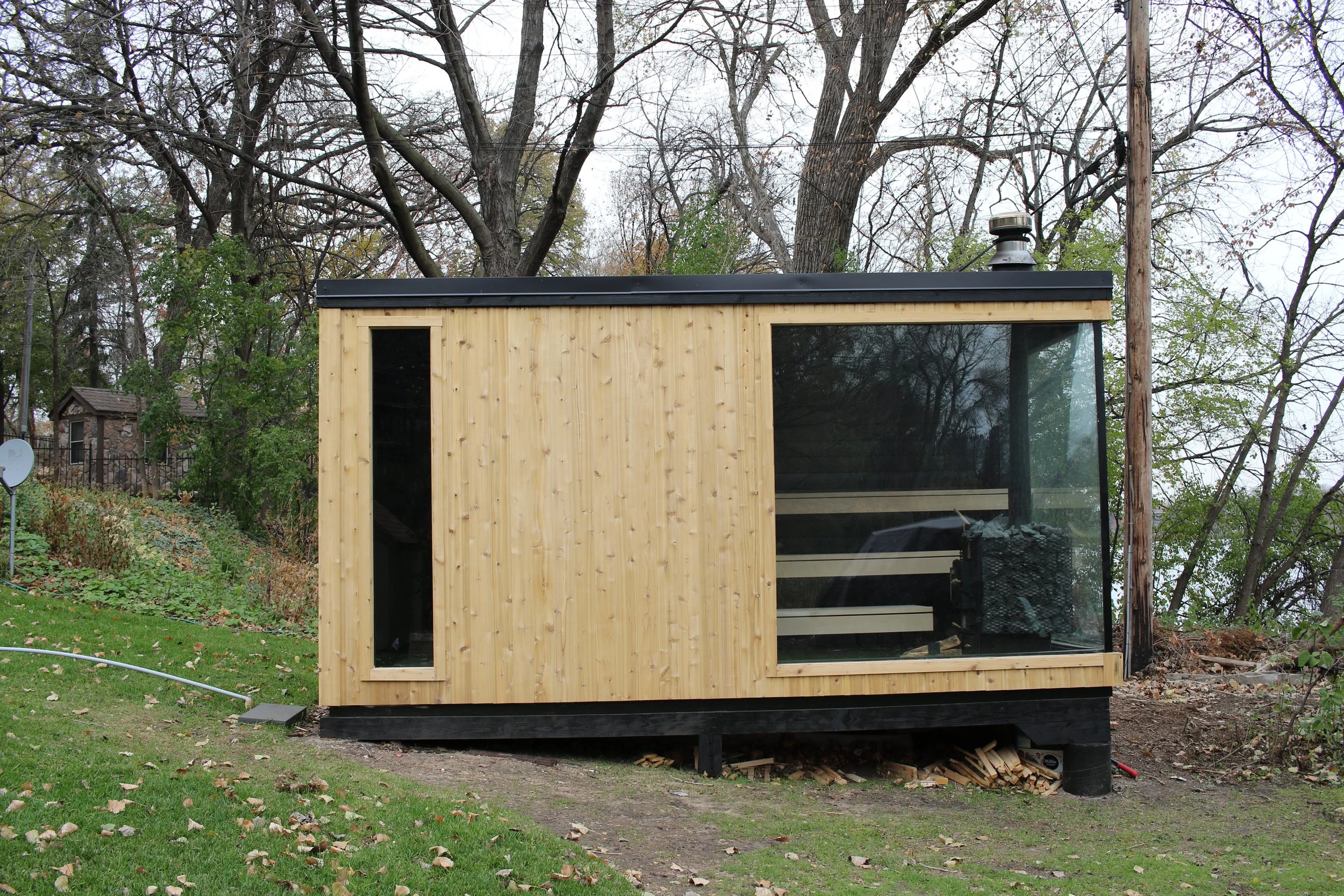 Small modern cabin with a large glass window and unfinished wooden siding, situated in a yard with trees and grass, under an overcast sky.