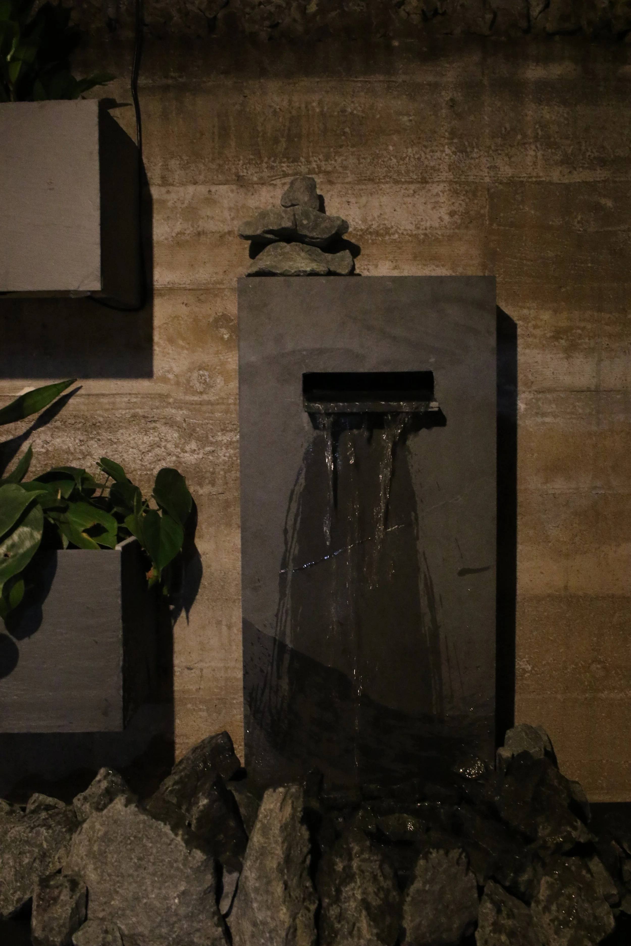 Indoor modern water fountain with a black rectangular plaque, with water flowing down its surface, surrounded by rocks at the base, against a textured brown wall, with a potted plant on the left.